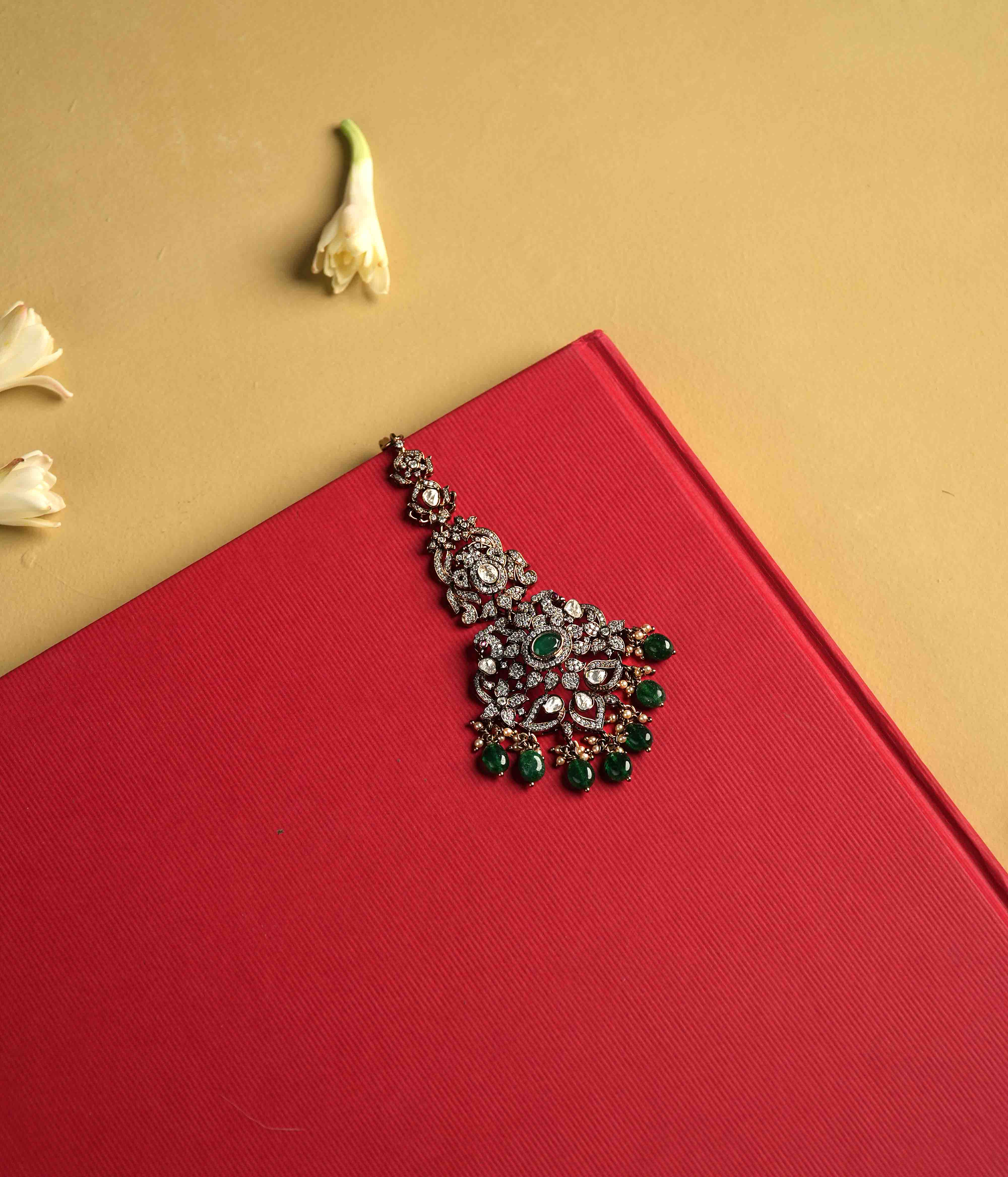 This intricately crafted Maangtika, handmade from 92.5 sterling silver with 14ct dark antique gold plating, features polki stones, CZ stones, green gemstones, green beads, and pearls.  The design shines with polki and moissanite brilliance, with cultured emerald dangles adding a touch of majestic elegance, evoking the opulence of a Victorian era.