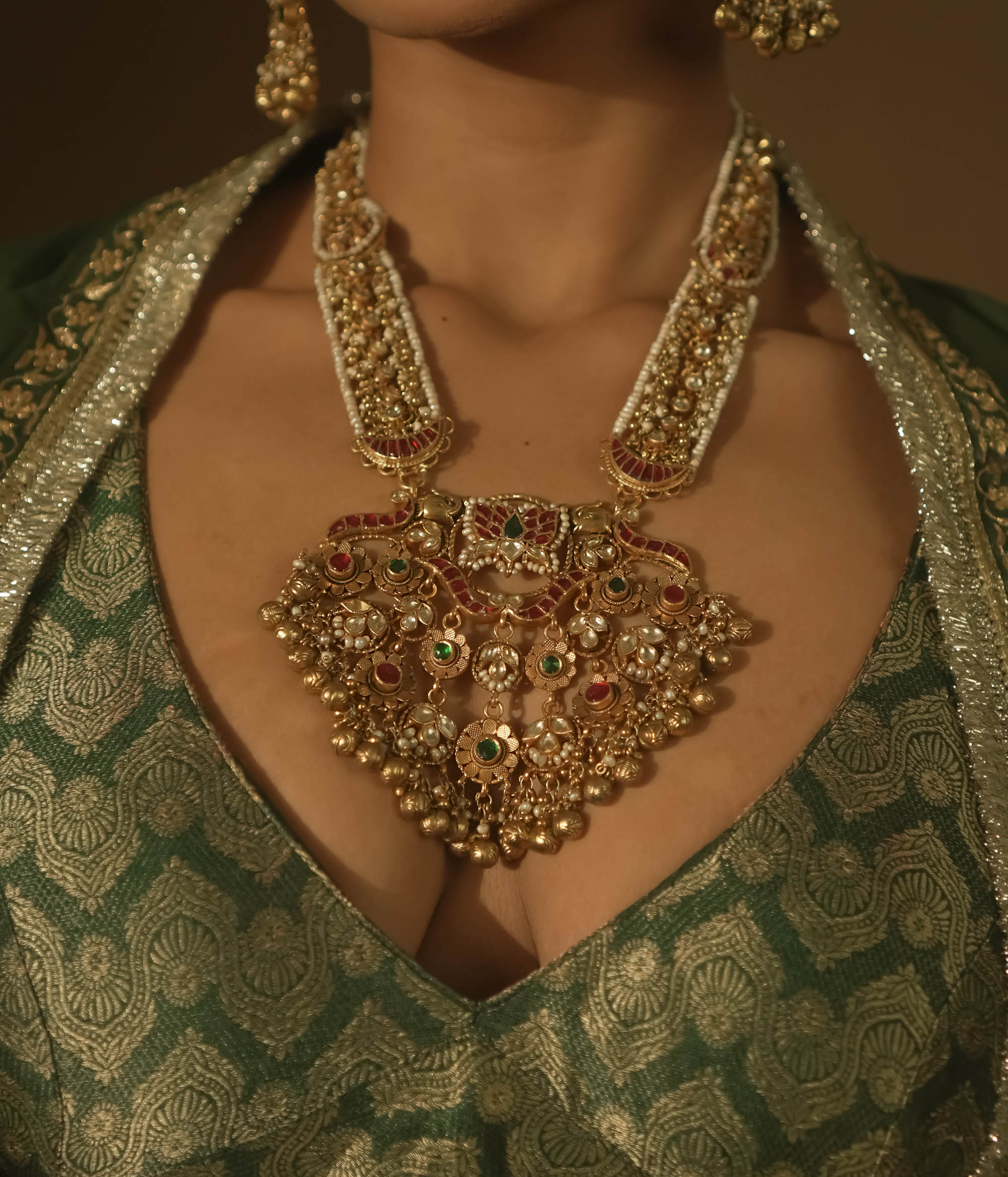 Gold-plated necklace with red and green Kundan stones, seed pearls, and dangling bells.