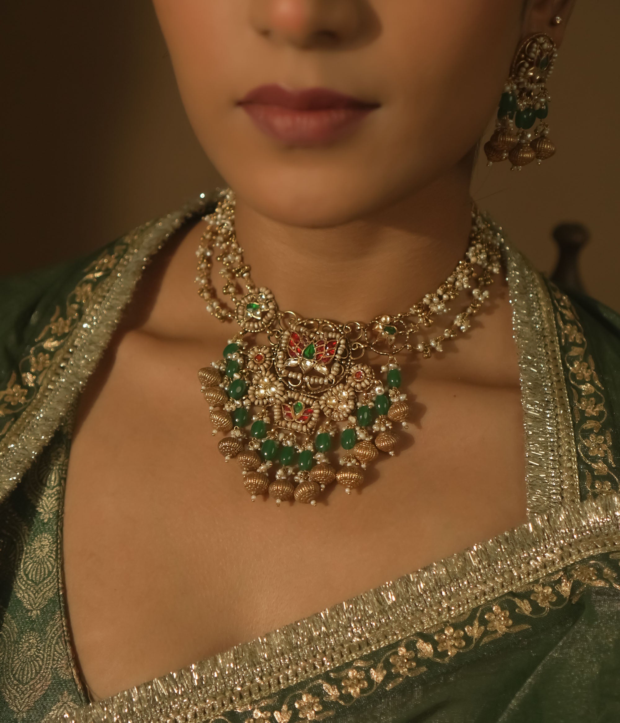 Gold necklace with green and red Kundan stones, beads, and a central pendant with bells.
