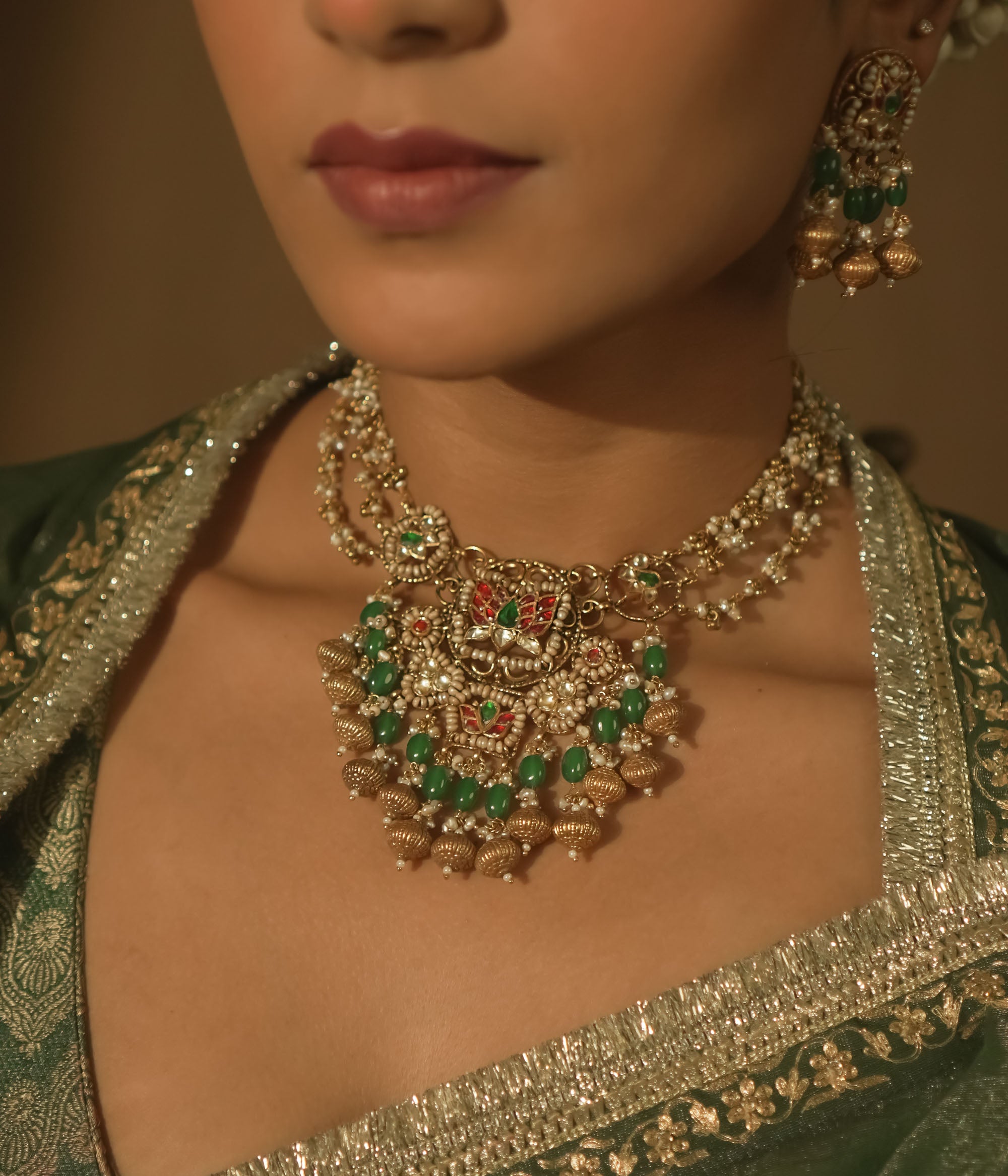 Gold necklace with green and red Kundan stones, beads, and a central pendant with bells.