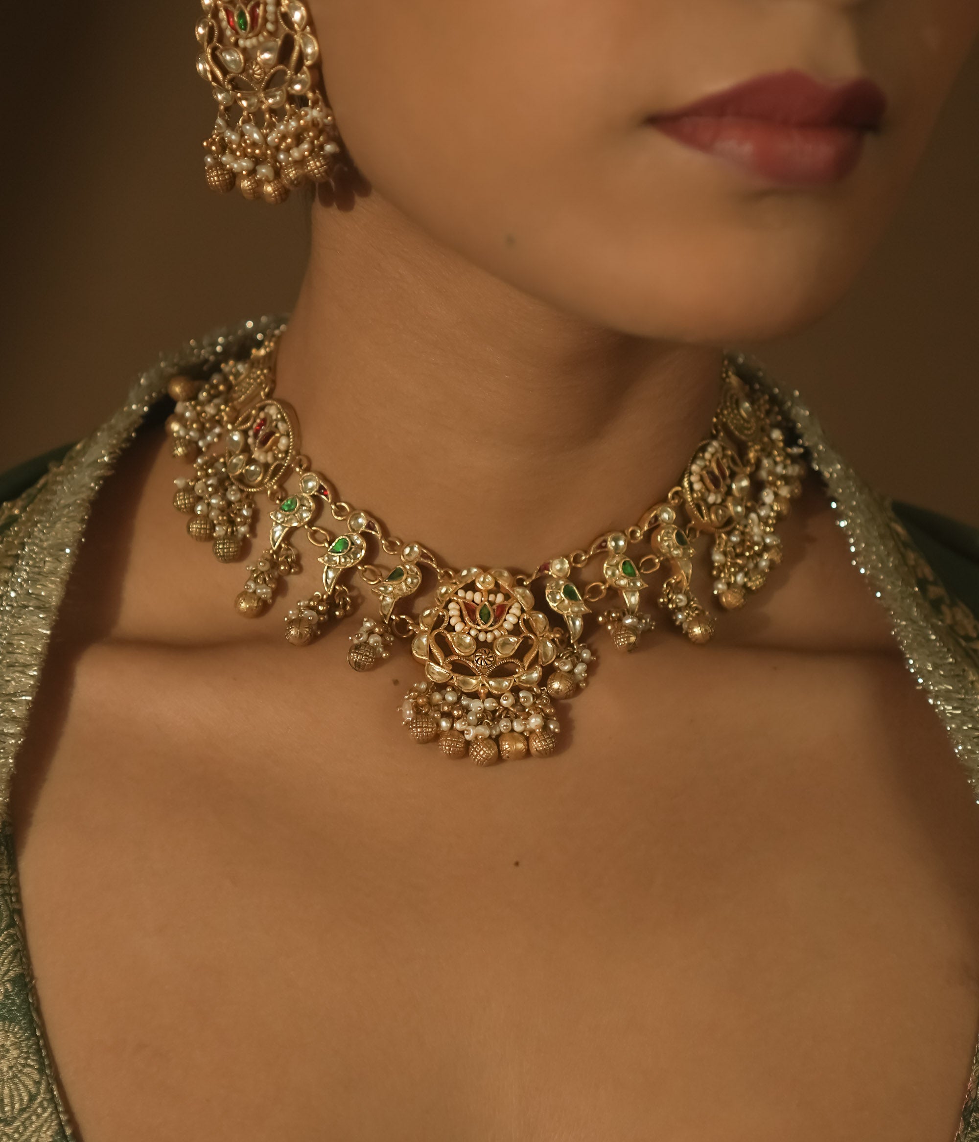 Intricate gold necklace with red and green Kundan stones, pearls, and parrot motifs.