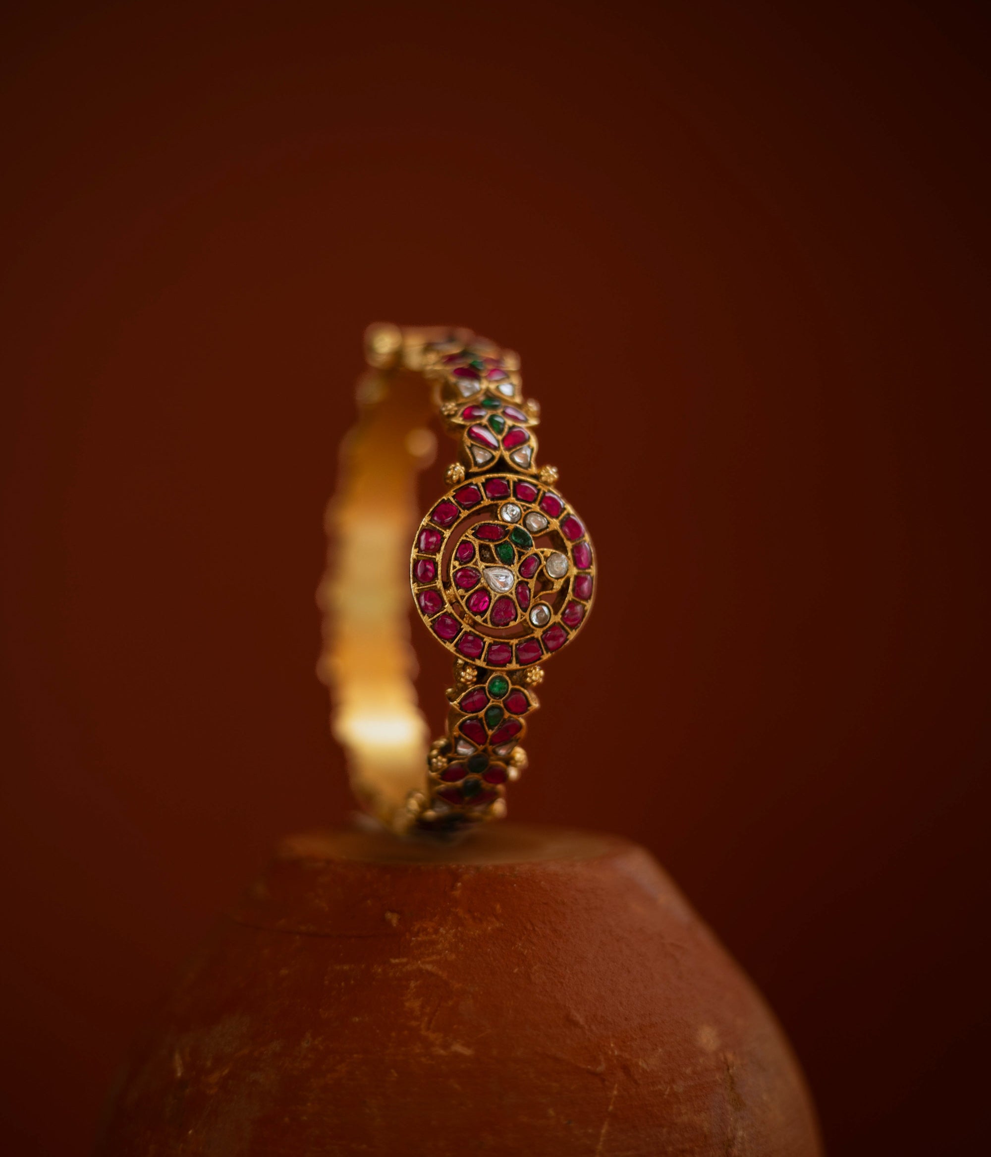 Gold bangle with circular kundan design, featuring red, green, and white stones in traditional pattern.