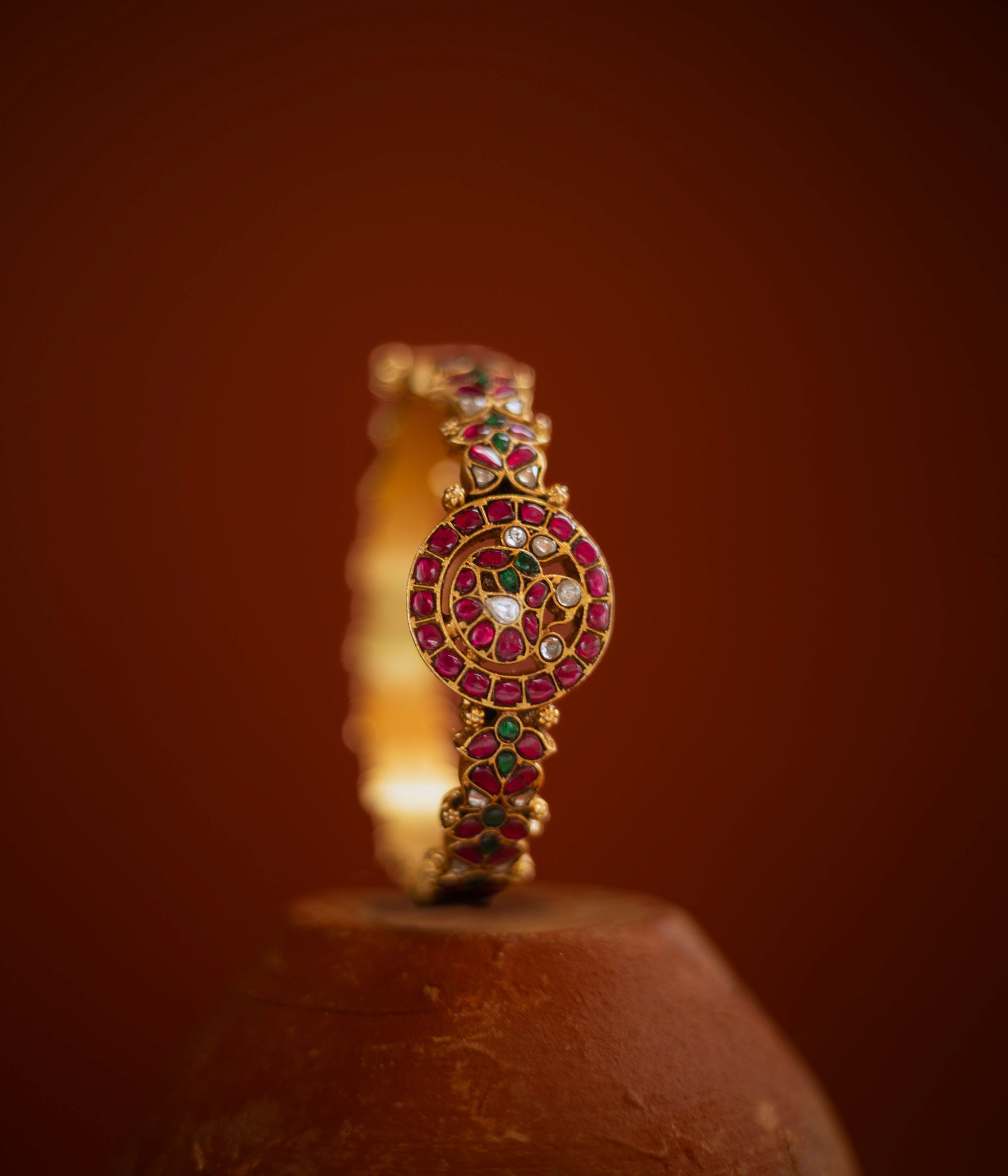 Gold bangle with circular kundan design, featuring red, green, and white stones in traditional pattern.