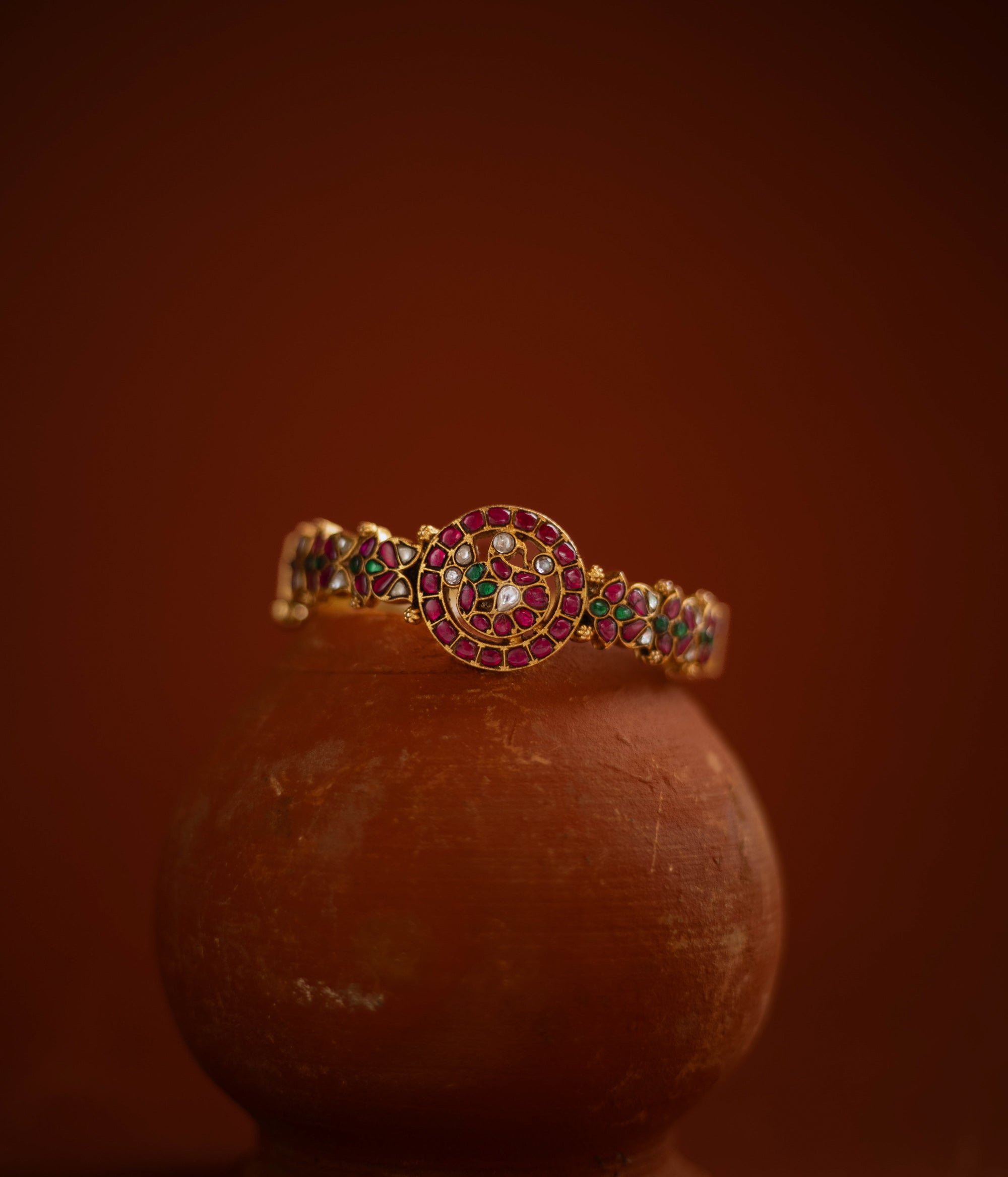 Gold bangle with circular kundan design, featuring red, green, and white stones in traditional pattern.