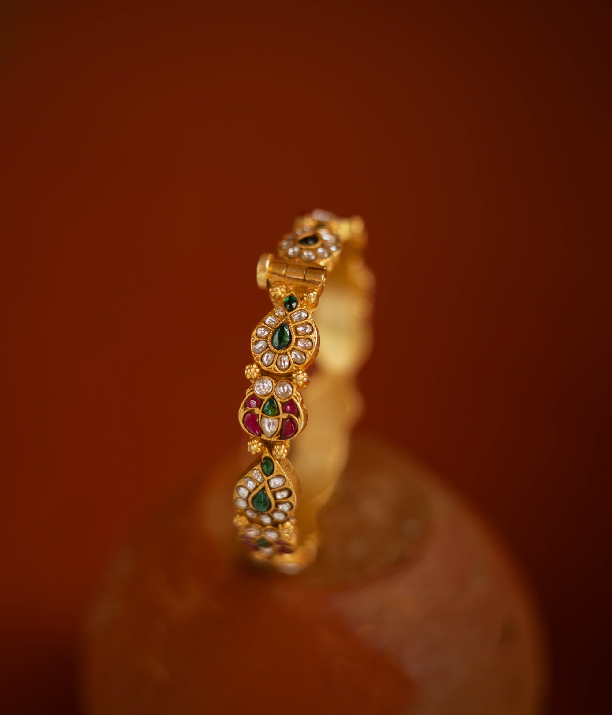 Gold bangle with red, green, and white kundan stones in traditional floral design.
