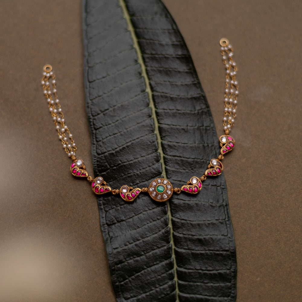 Gold-toned necklace with red, white, and green kundan stones and pearl chains, displayed on a dark green leaf.