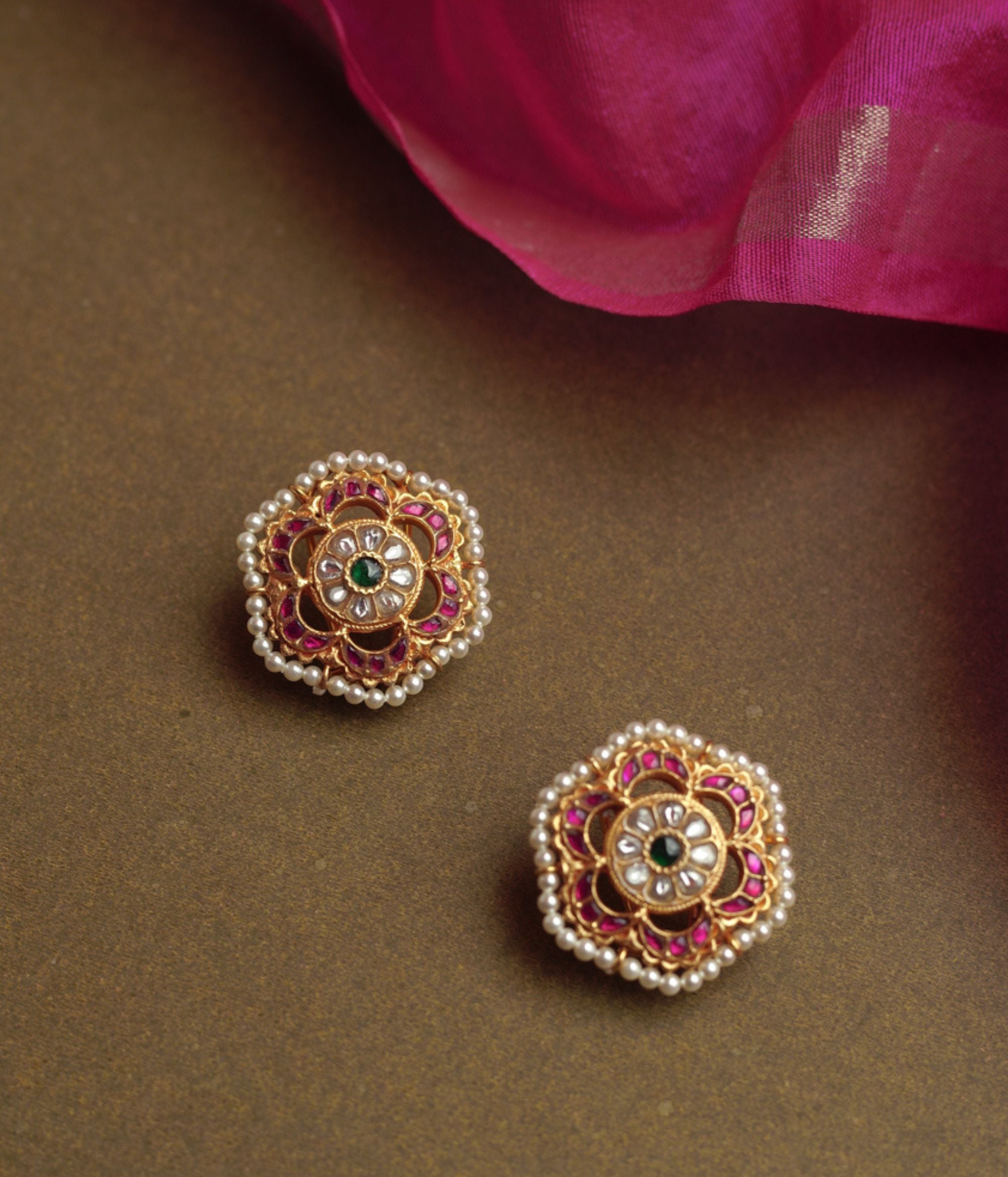 A pair of round gold stud earrings with a floral design. Each features a central green stone, surrounded by white stones, an outer ring of red stones, and a fringe of pearls.