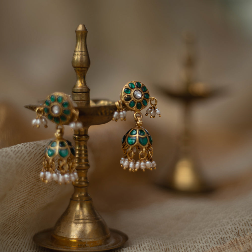 Gold jhumka earrings with green kundan stones and pearl drops, displayed on a brass oil lamp.