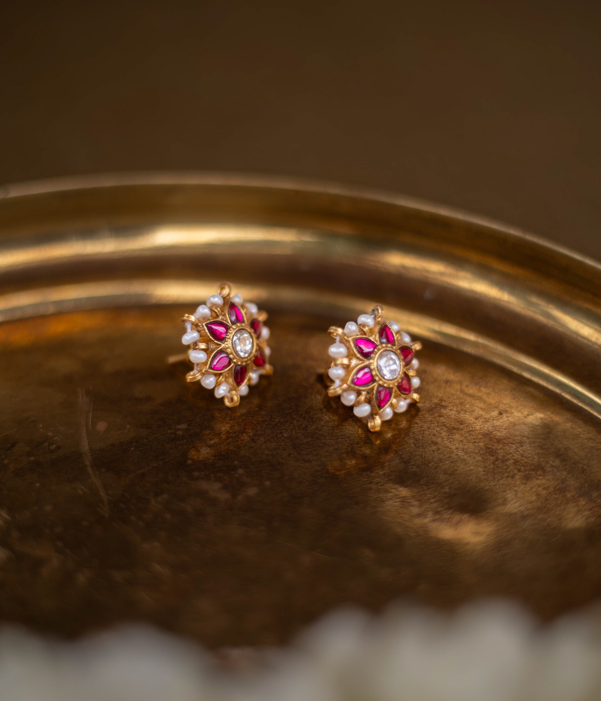 Meira Kundan Earrings with red and white kundan stones and pearl accents on a floral base, placed on a golden brass surface.