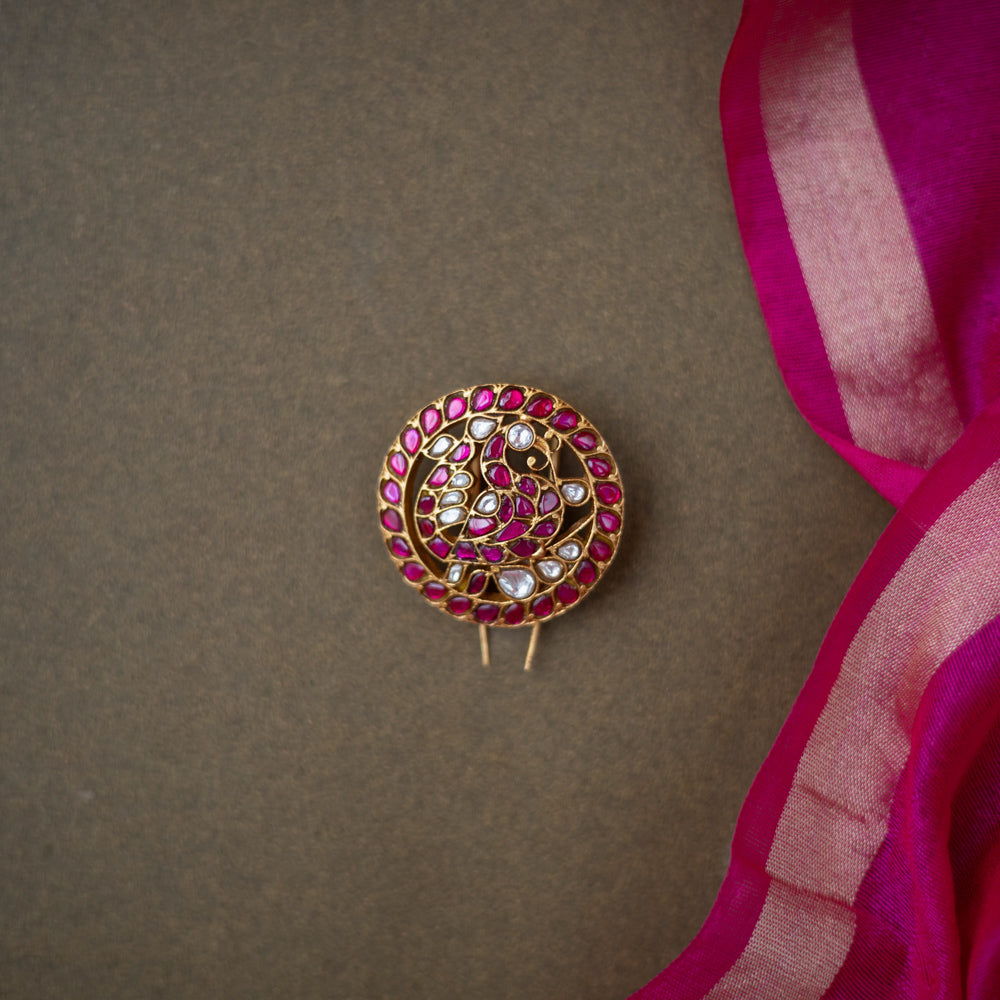 Round paisley-motif kundan hairpin with red and white stones, placed on a dark brown surface with a pink silk saree.