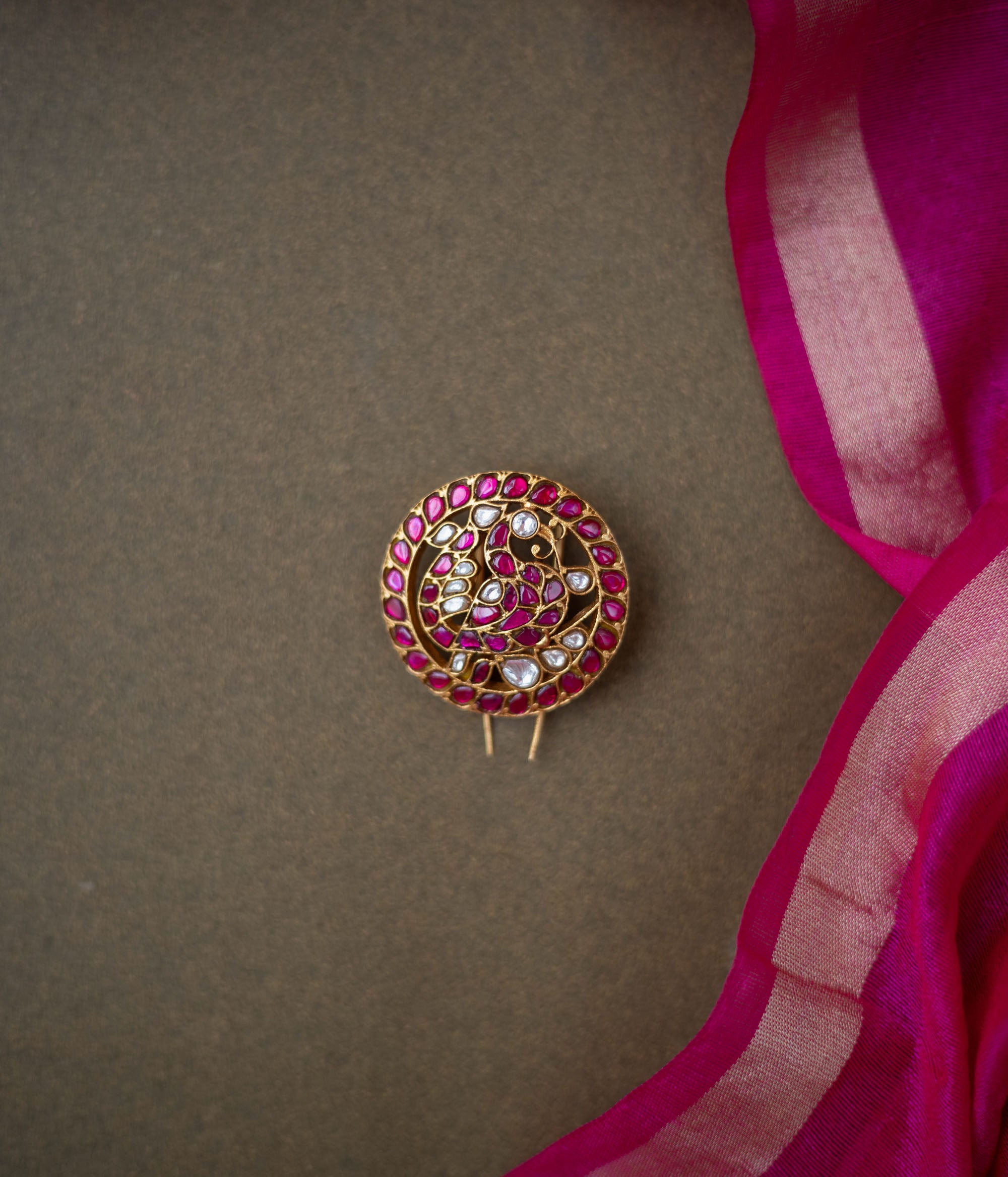 Round paisley-motif kundan hairpin with red and white stones, placed on a dark brown surface with a pink silk saree.