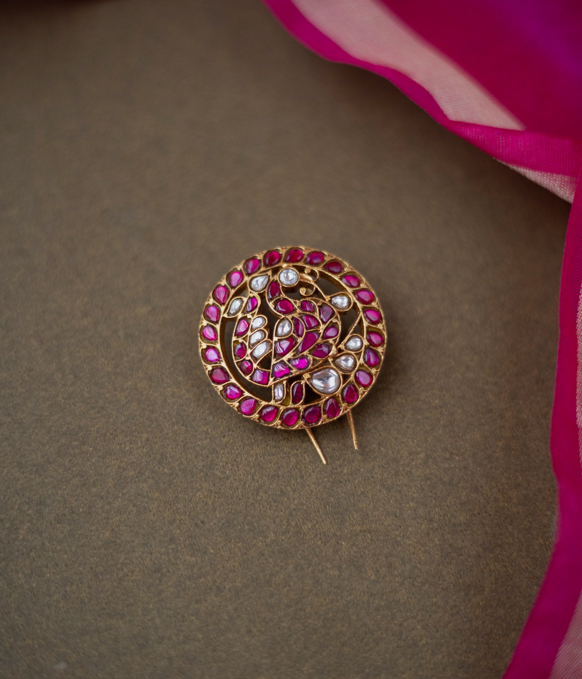 Round paisley-motif kundan hairpin with red and white stones, placed on a dark brown surface with a pink silk saree.