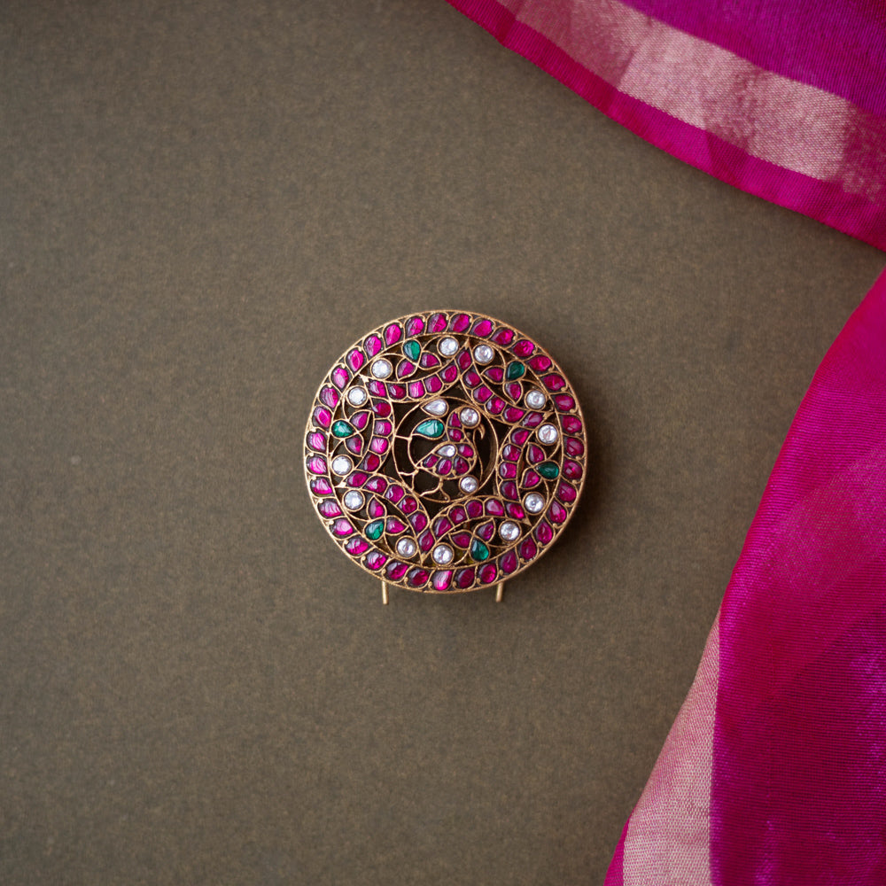 Round peacock-themed kundan hairpin with red, green, and white stones on a dark surface with a pink silk saree border.