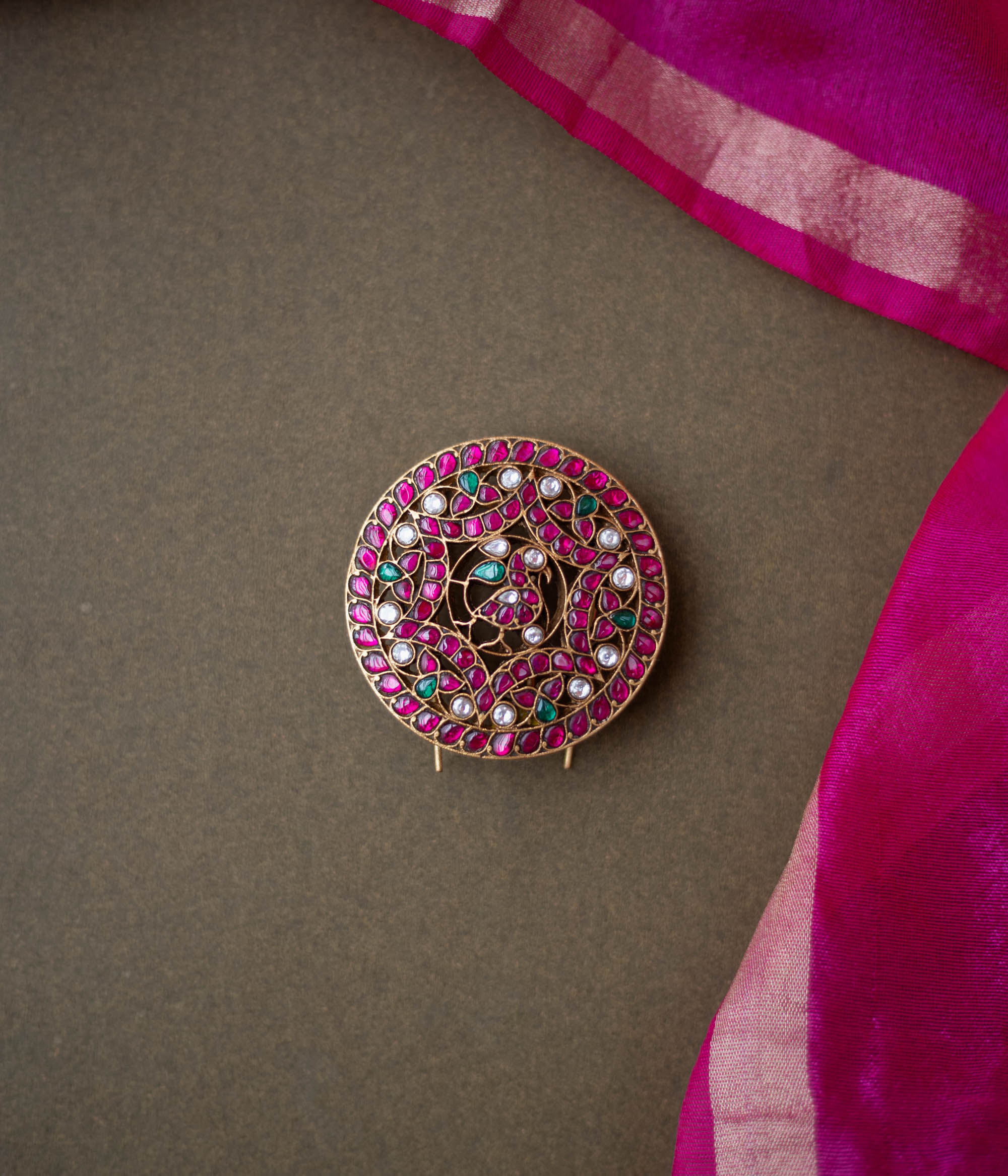 Round peacock-themed kundan hairpin with red, green, and white stones on a dark surface with a pink silk saree border.