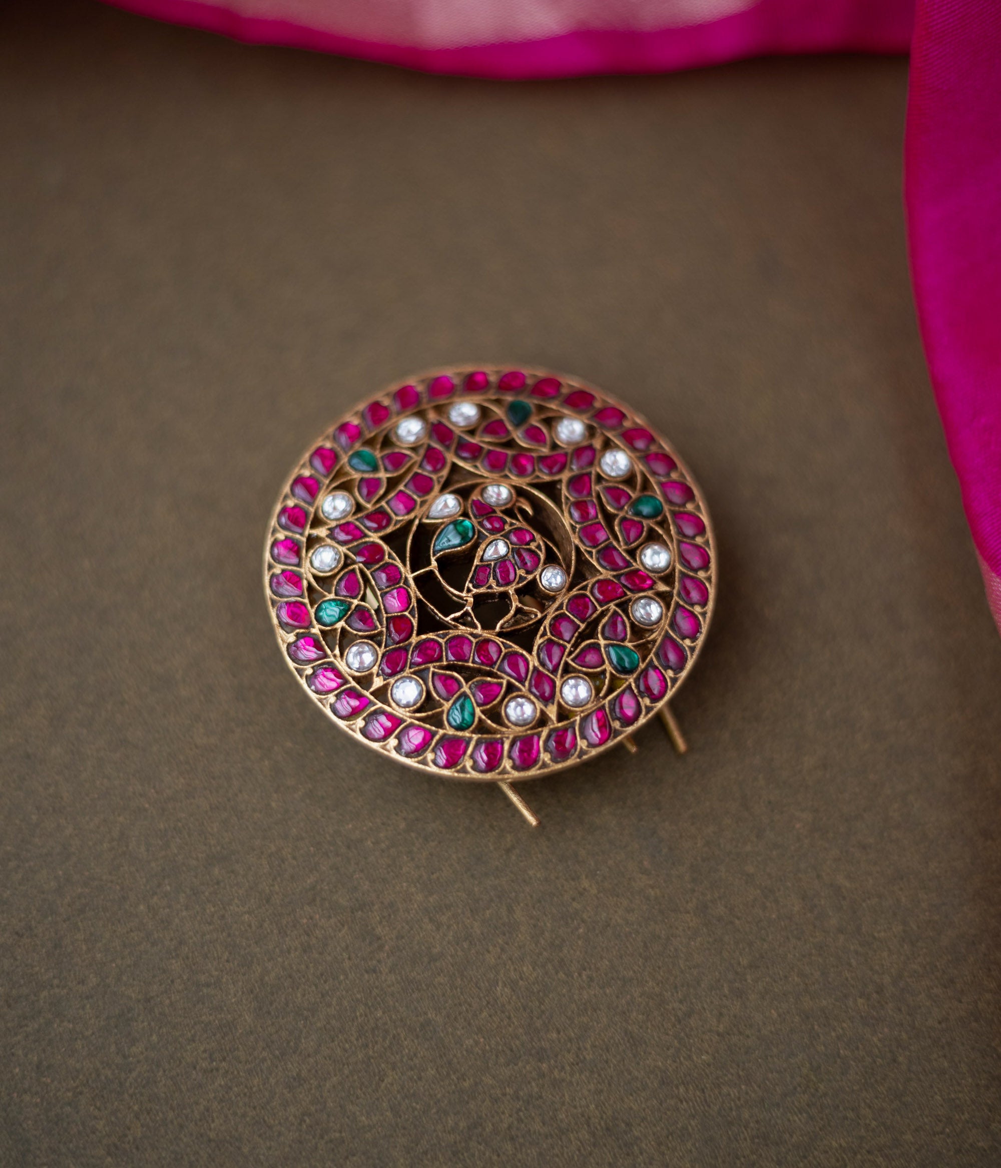 Round peacock-themed kundan hairpin with red, green, and white stones on a dark surface with a pink silk saree border.
