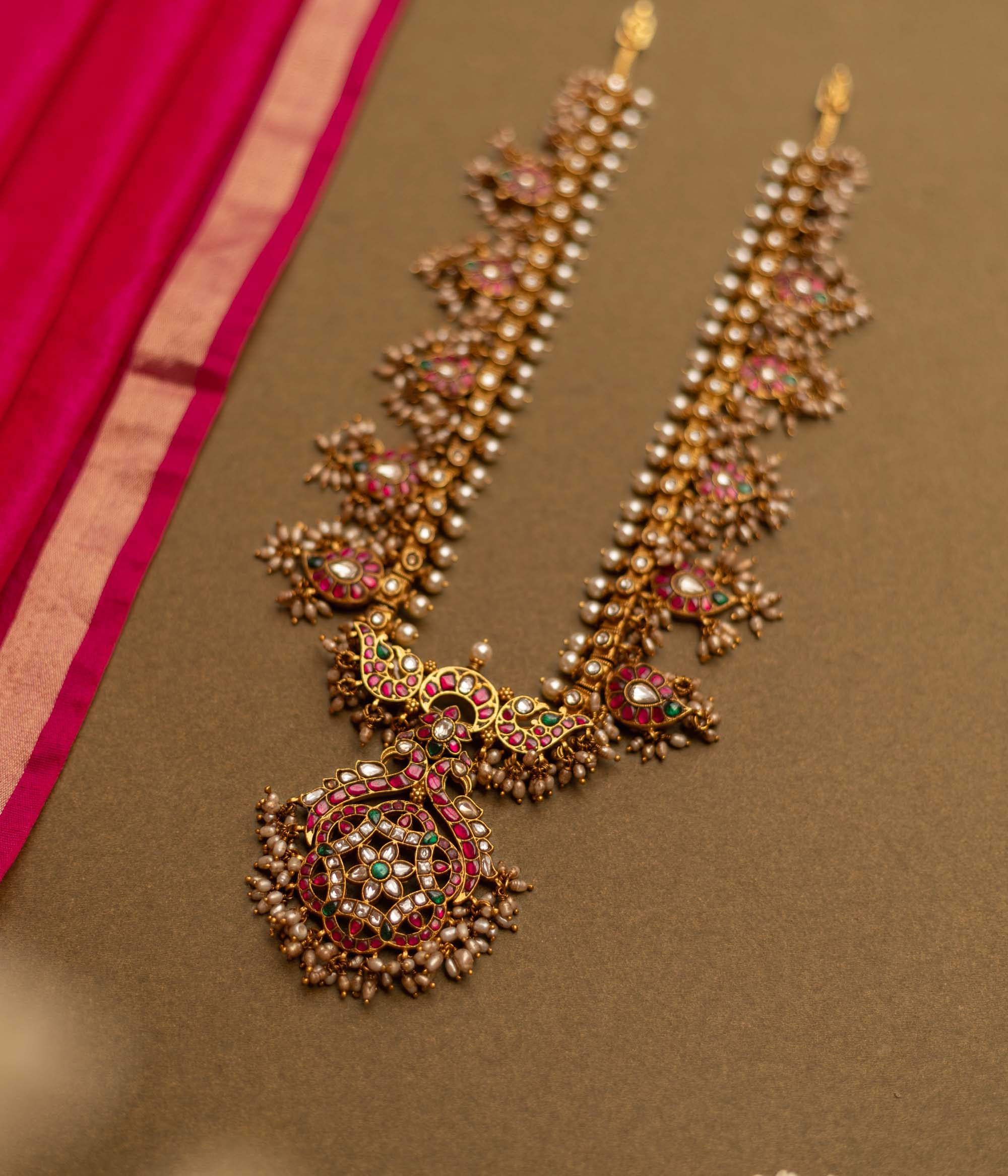Overhead view of a Gutta Pusalu necklace with pearls and colored stones. The necklace rests on a brown surface with a pink fabric accent.