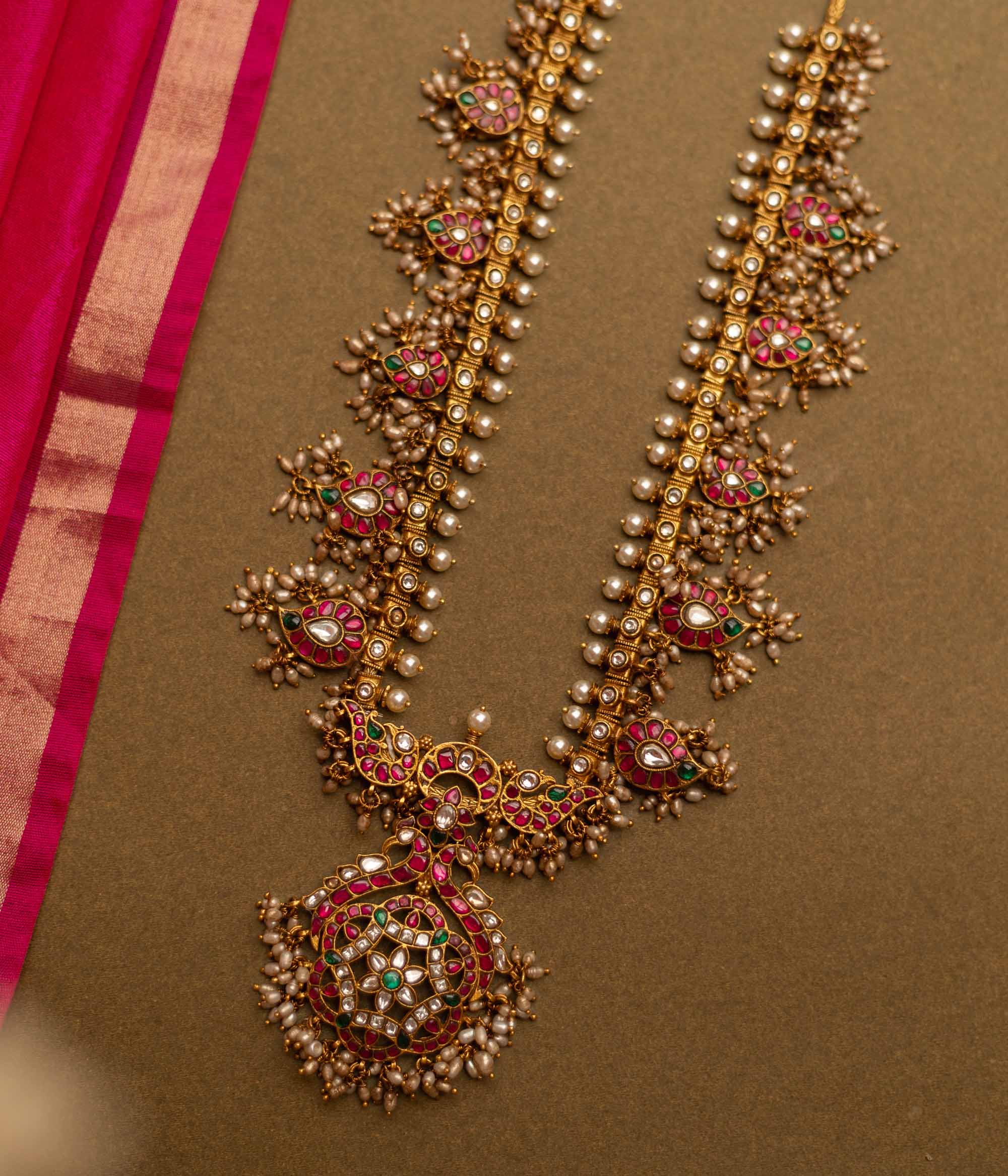 Overhead view of a Gutta Pusalu necklace with pearls and colored stones. The necklace rests on a brown surface with a pink fabric accent.