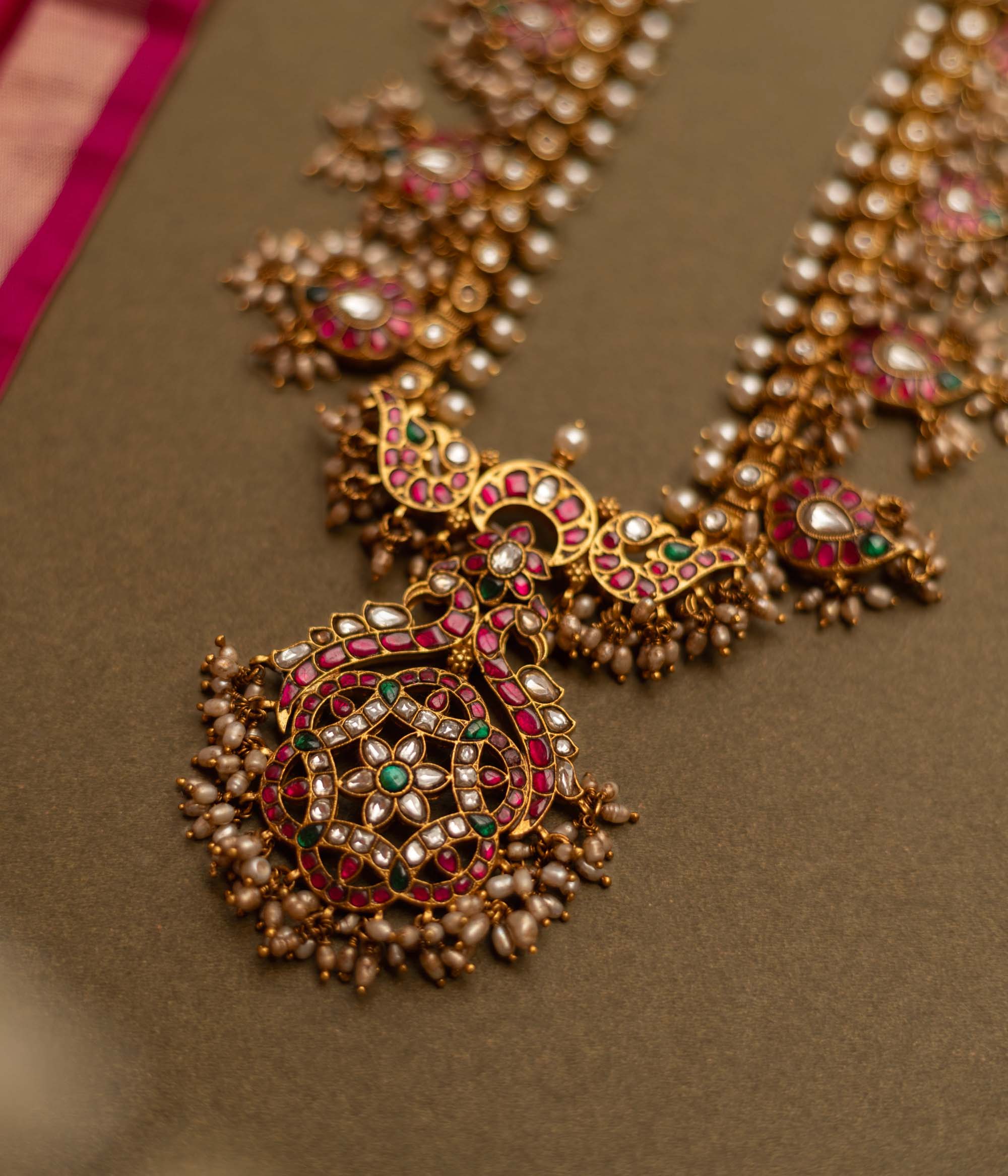 Overhead view of a Gutta Pusalu necklace with pearls and colored stones. The necklace rests on a brown surface with a pink fabric accent.