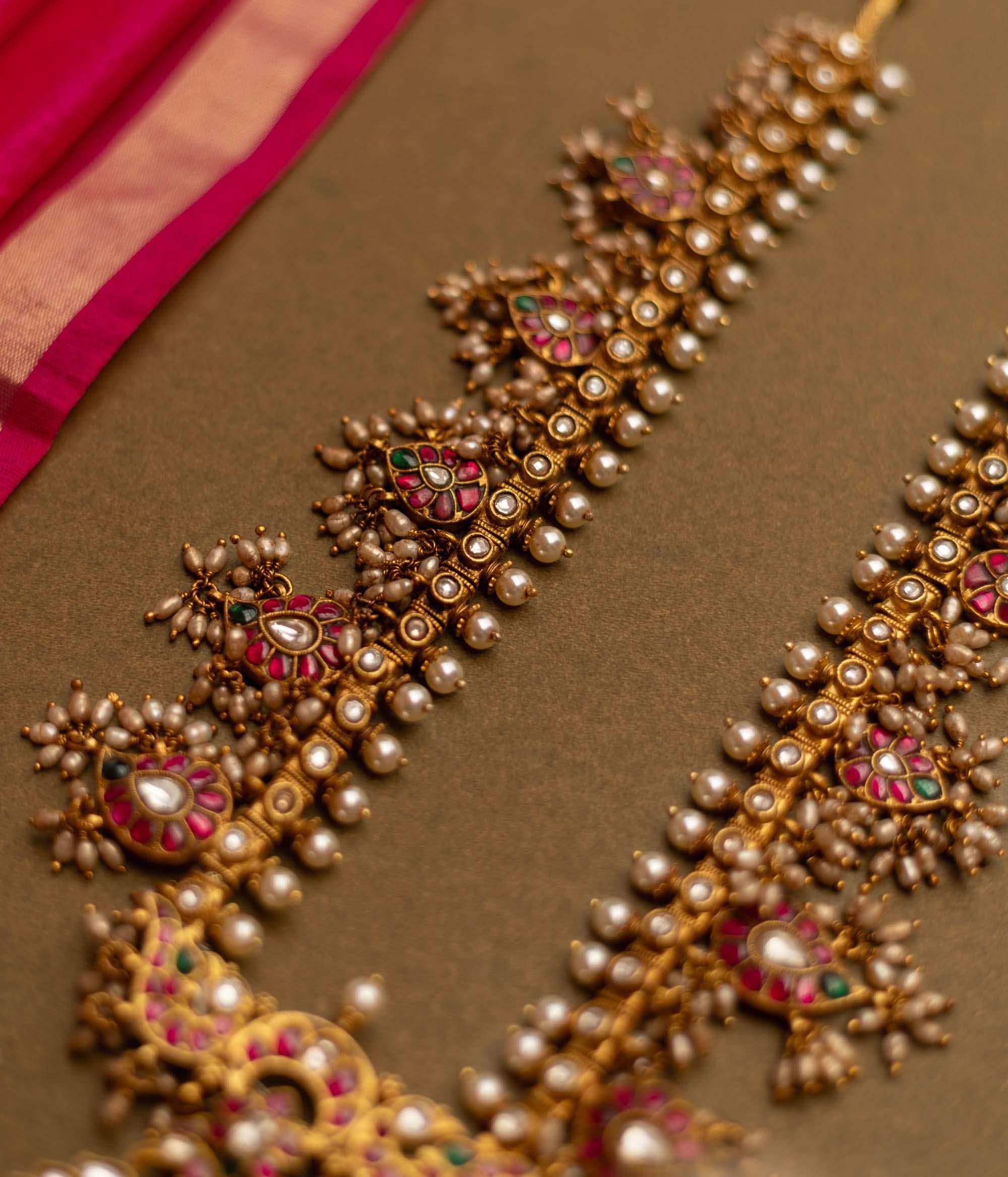 Overhead view of a Gutta Pusalu necklace with pearls and colored stones. The necklace rests on a brown surface with a pink fabric accent.
