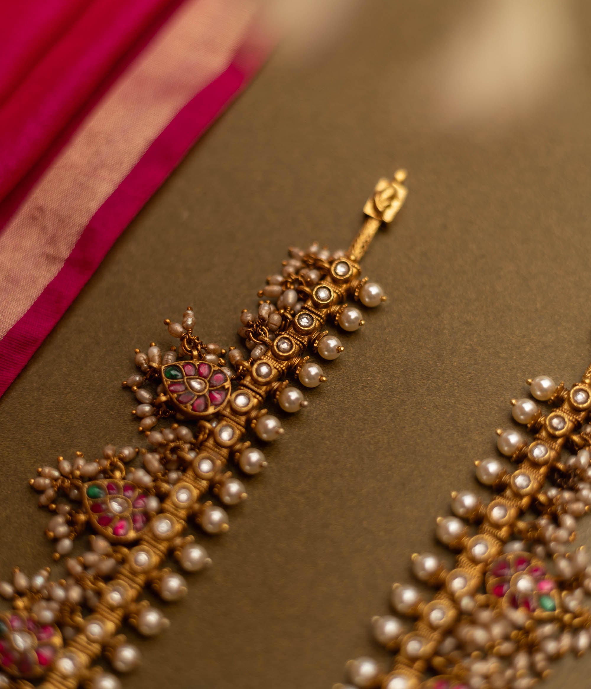 Overhead view of a Gutta Pusalu necklace with pearls and colored stones. The necklace rests on a brown surface with a pink fabric accent.