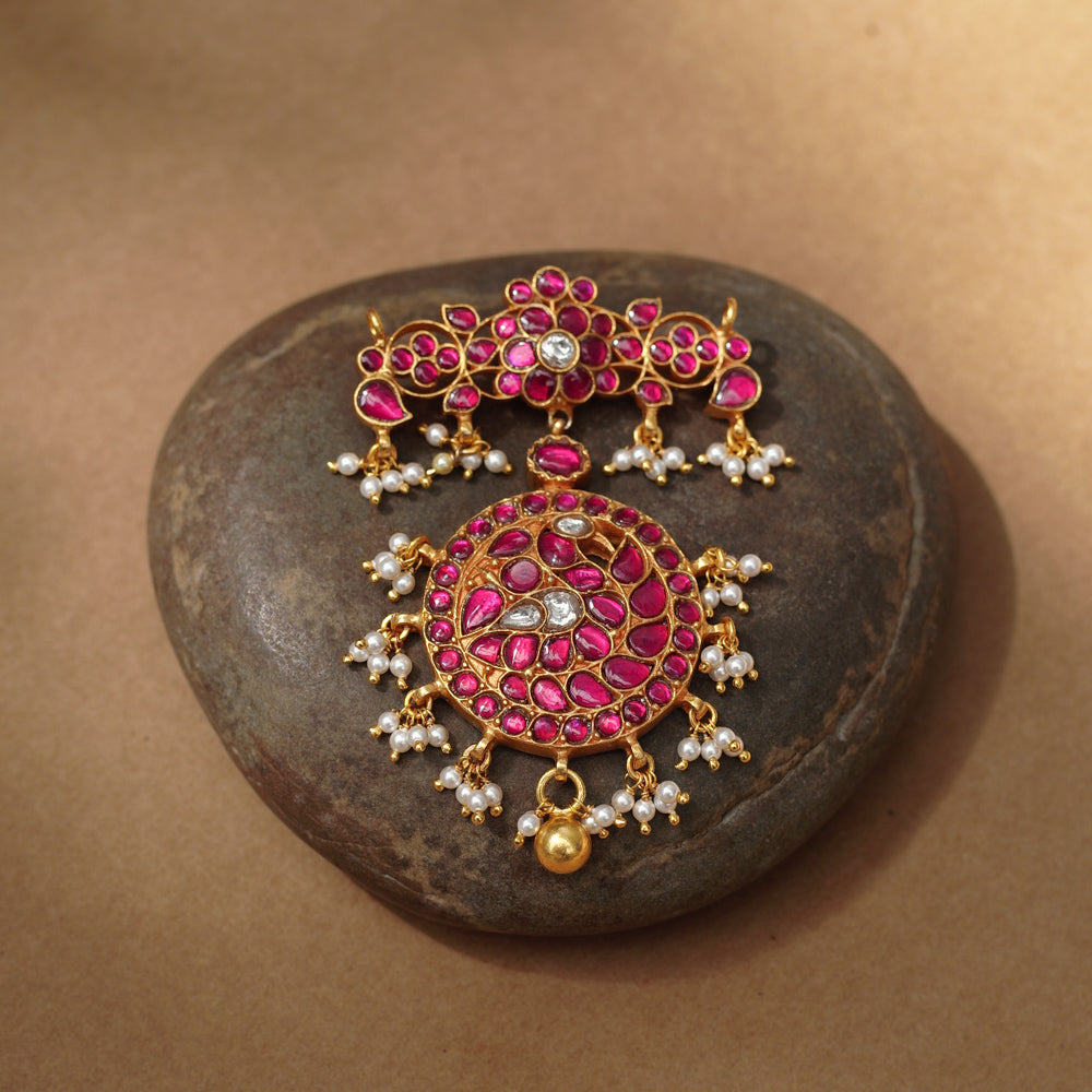 Close-up of a large gold Kundan pendant. It features a circular main section and a floral top piece, both adorned with red and white Kundan stones and dangling pearls.