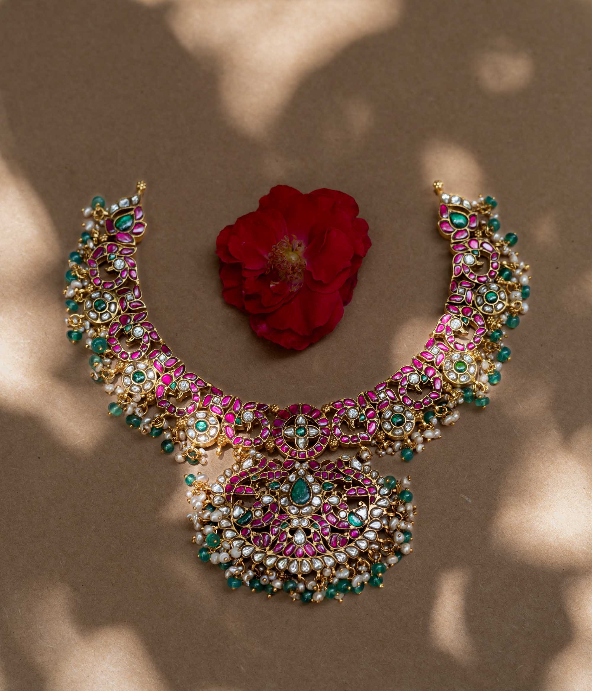 A handcrafted 22ct gold-plated 92.5 sterling silver Padmavati Kemp necklace, featuring red, green, and white Kundan stones, green beads, and pearls, with a broad, ornate design and a central pearl-clustered pendant, ideal for bridal wear and grand celebrations.