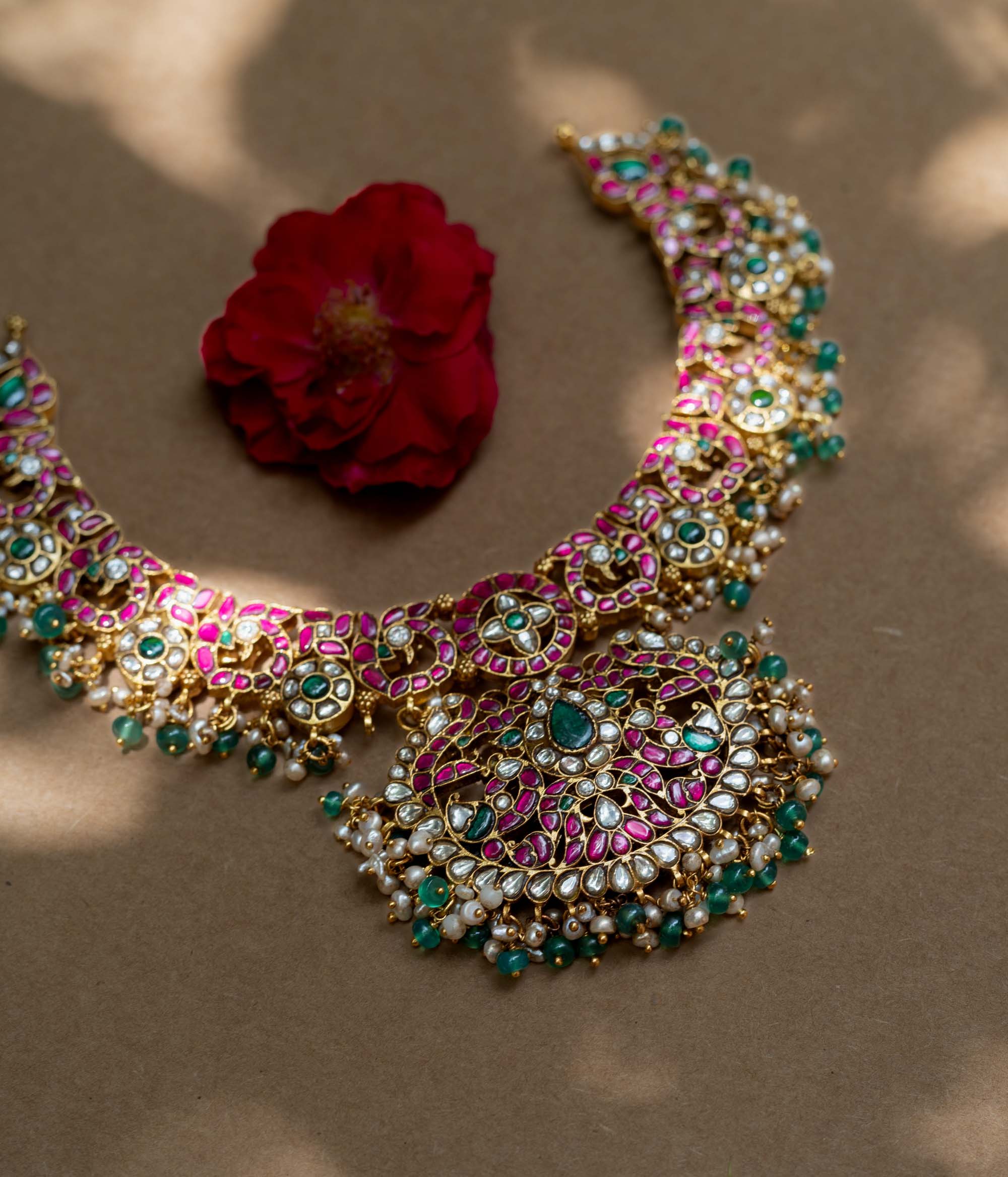 A handcrafted 22ct gold-plated 92.5 sterling silver Padmavati Kemp necklace, featuring red, green, and white Kundan stones, green beads, and pearls, with a broad, ornate design and a central pearl-clustered pendant, ideal for bridal wear and grand celebrations.