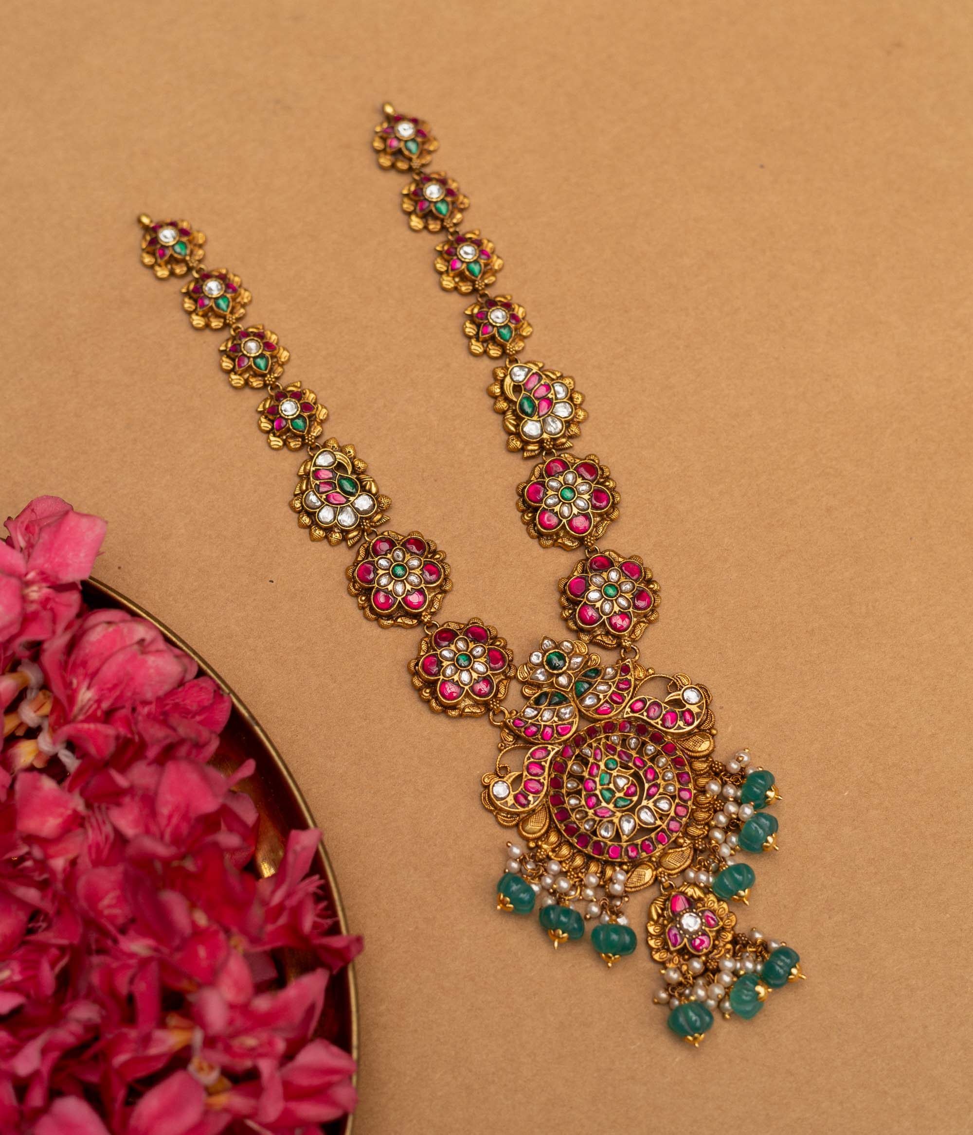 A 92.5 sterling silver Blossom Radiance Peacock Necklace with an antique finish, featuring a pendant adorned with twin peacocks, dazzling kundan stones, and vibrant pink and green accents, a floral-link chain, and dangling emerald-green beads and pearls, perfect for weddings, temple occasions, and cultural festivities.