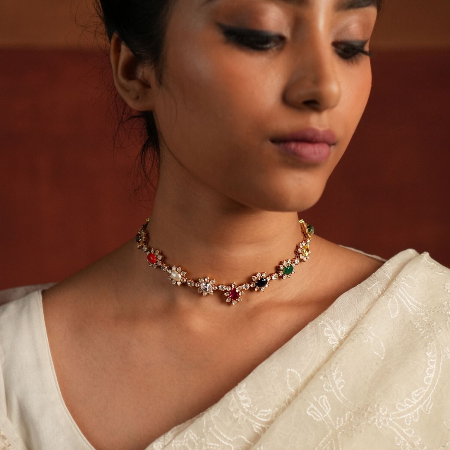 A sophisticated gold-plated silver choker featuring a delicate chain adorned with exquisite Navratna stones arranged in a charming floral design. Each vibrant gemstone is beautifully encased by shimmering CZ stones, adding a touch of elegance and brilliance. This timeless piece seamlessly blends tradition with modern sophistication, making it a perfect accessory for any occasion.
