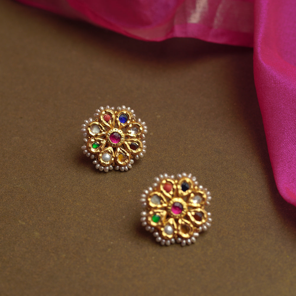 Close-up of two ornate gold stud earrings. Each features a central red gem surrounded by colorful Navratna stones and an outer border of small pearls, resting on a brown surface.
