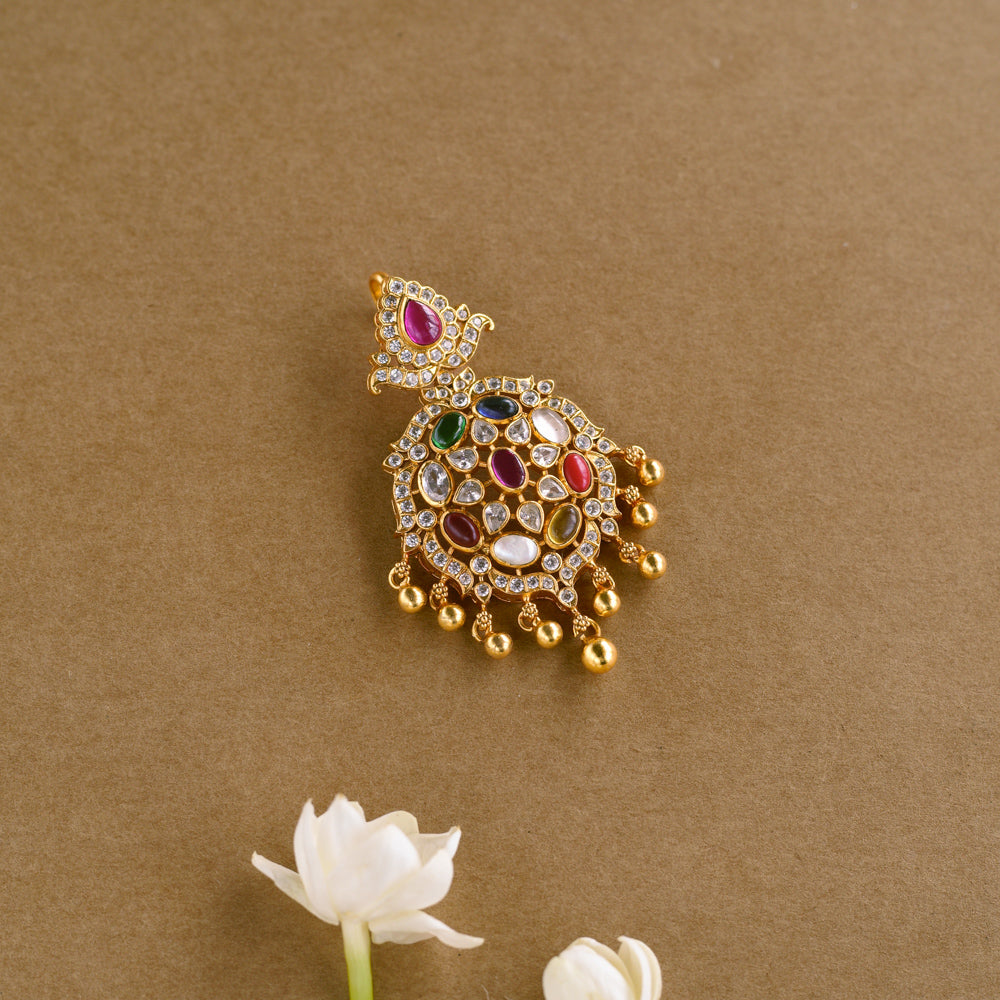 A gold pendant with an ornate, asymmetrical design. It is adorned with various colorful Navratna stones, clear stones, white Polki-like stones, and a single red stone at the top. Small gold beads dangle from the bottom.
