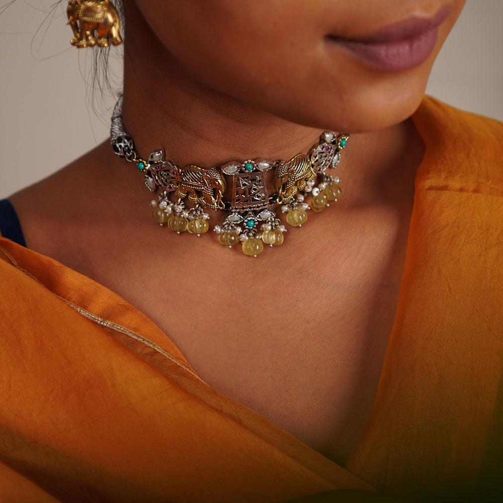 This oxidized silver choker, adorned with white, red, and green kundan stones and delicate pearl droplets, features regal gold-plated elephant motifs at its heart. A timeless piece that evokes the grandeur of a bygone era, reminiscent of a treasured heirloom.