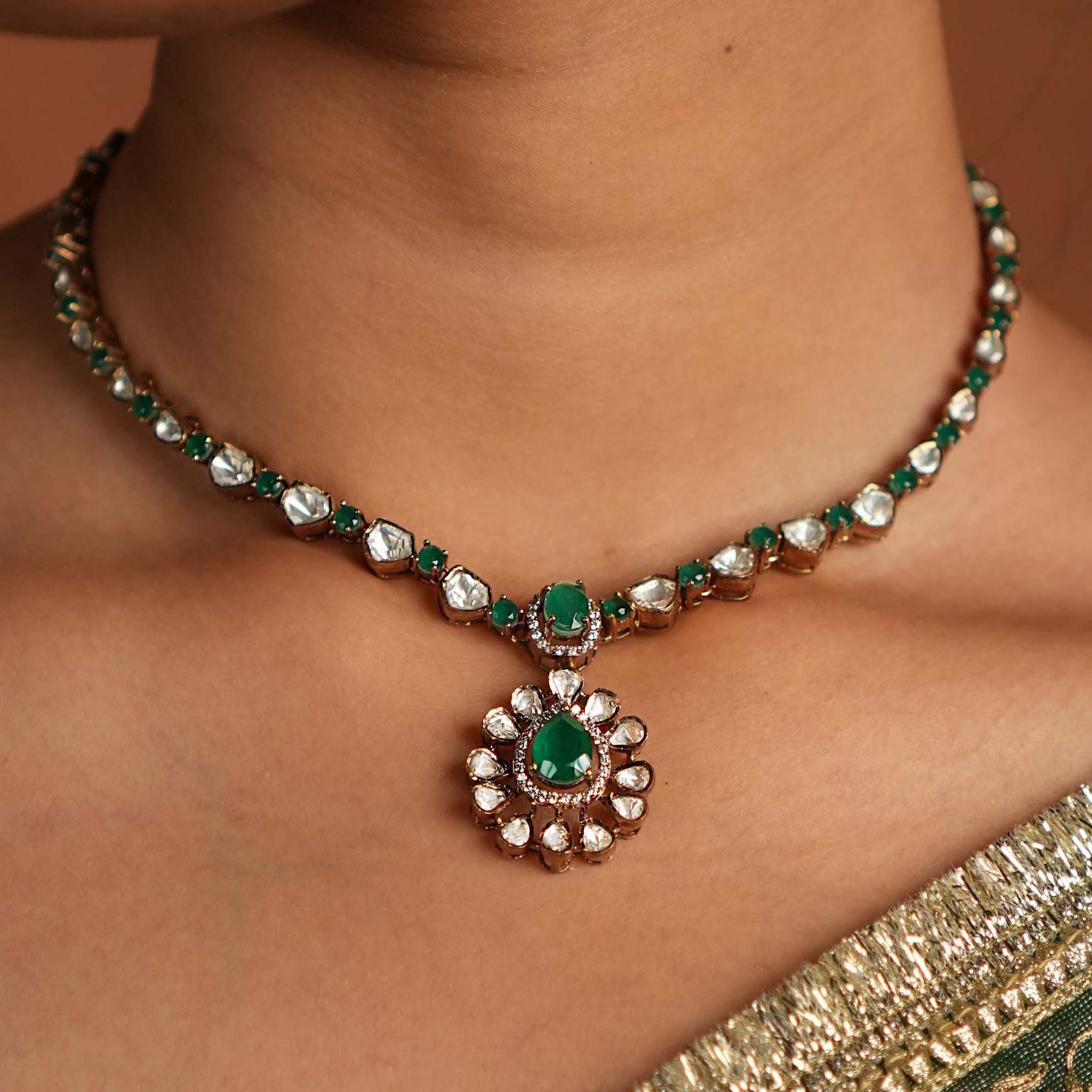 Close-up of a model wearing a gold-toned necklace with emerald-green stones, featuring a teardrop green pendant surrounded by sparkling stones, styled with a traditional outfit.