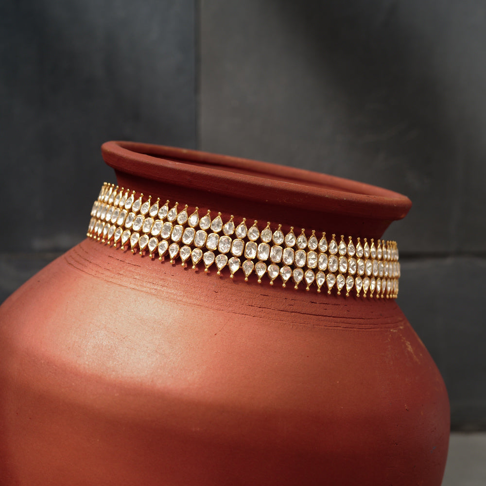 Close-up of a gold choker with three rows of teardrop-shaped white Polki stones. The choker is draped around a terracotta pot.