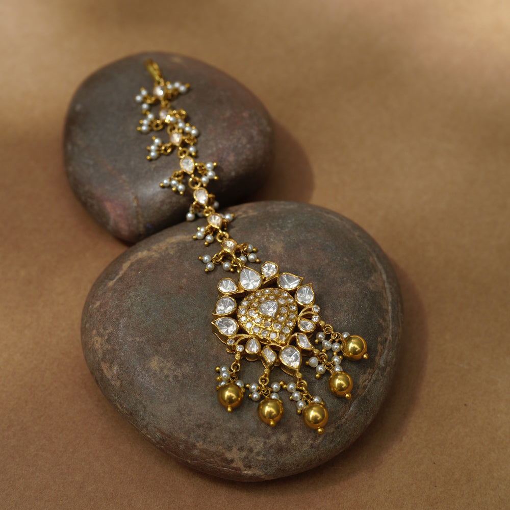 A golden maang tikka with a central heart-shaped pendant. It's adorned with white Polki stones and small dangling pearls, resting on two dark stones.
