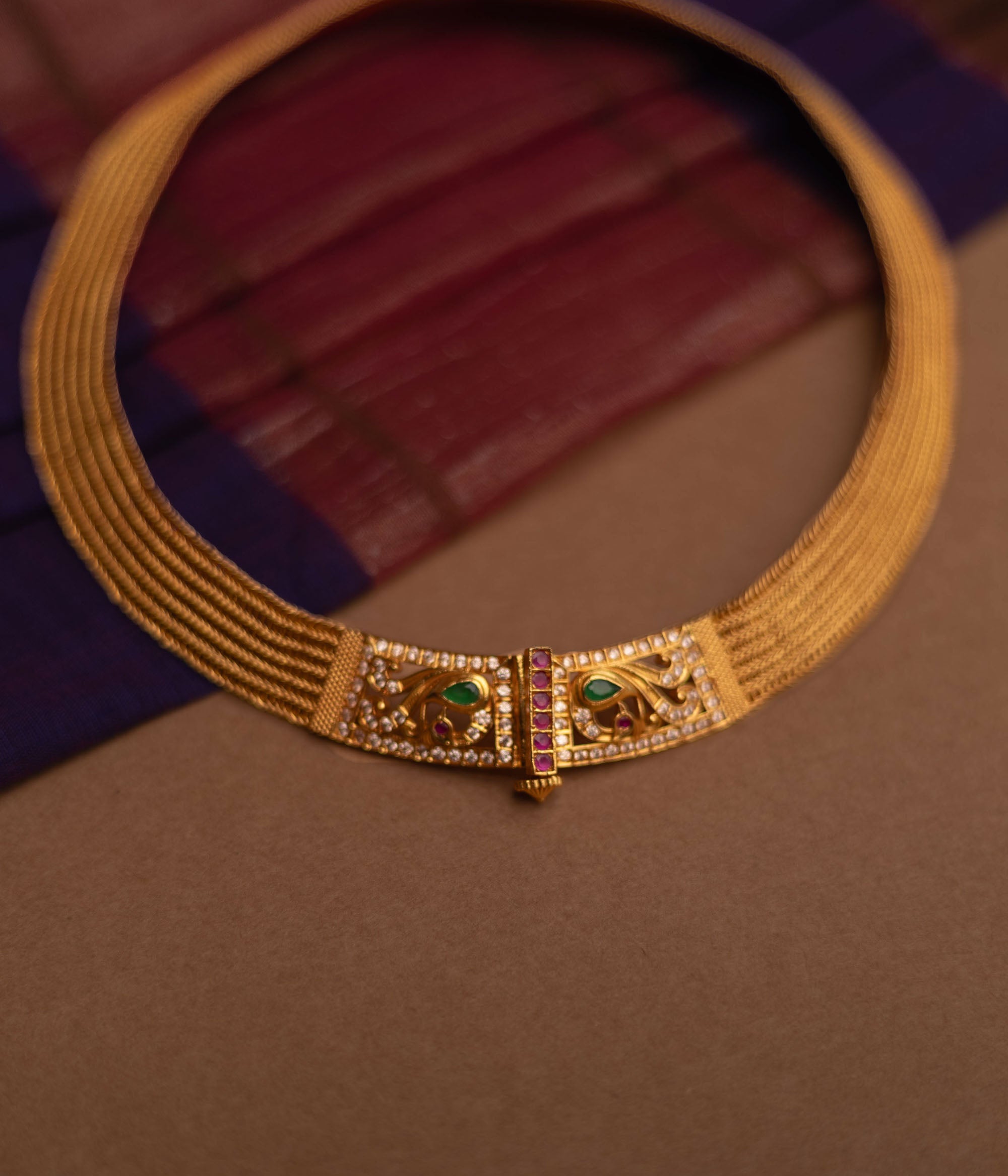 Close-up of a thick, golden choker necklace. It features intricate detailing with small green and clear stones at the clasp, set against a blurred fabric background.