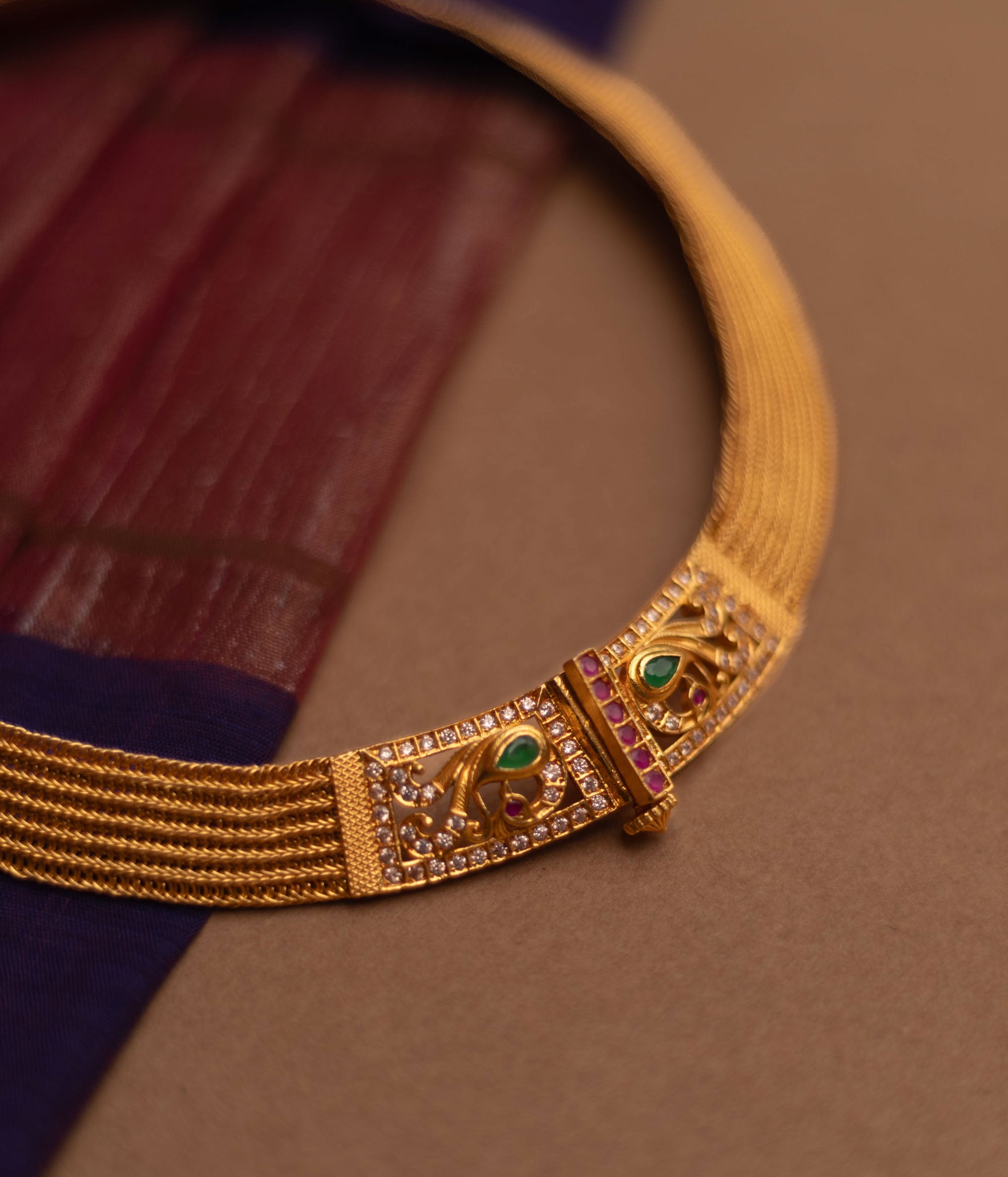 Close-up of a thick, golden choker necklace. It features intricate detailing with small green and clear stones at the clasp, set against a blurred fabric background.