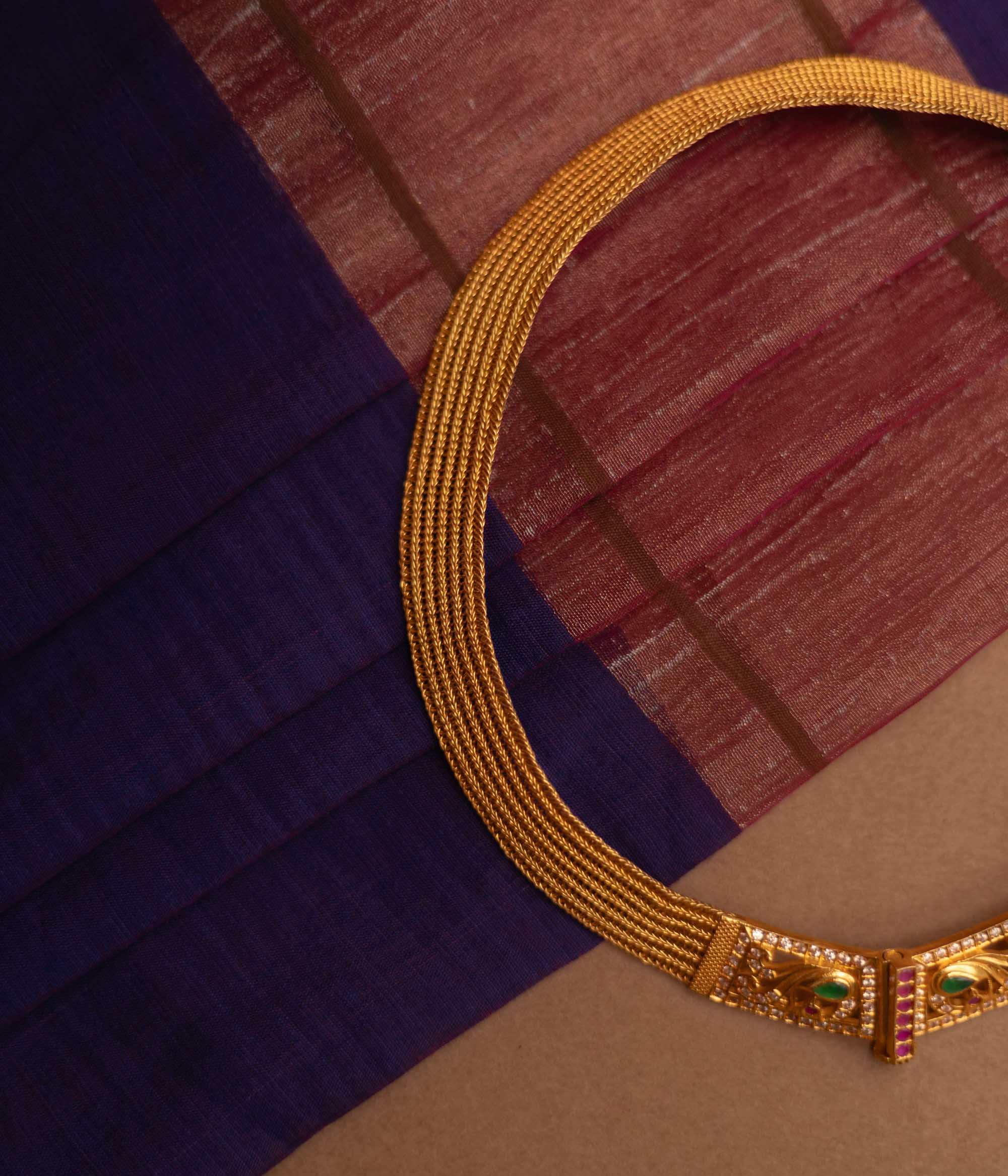 Close-up of a thick, golden choker necklace. It features intricate detailing with small green and clear stones at the clasp, set against a blurred fabric background.
