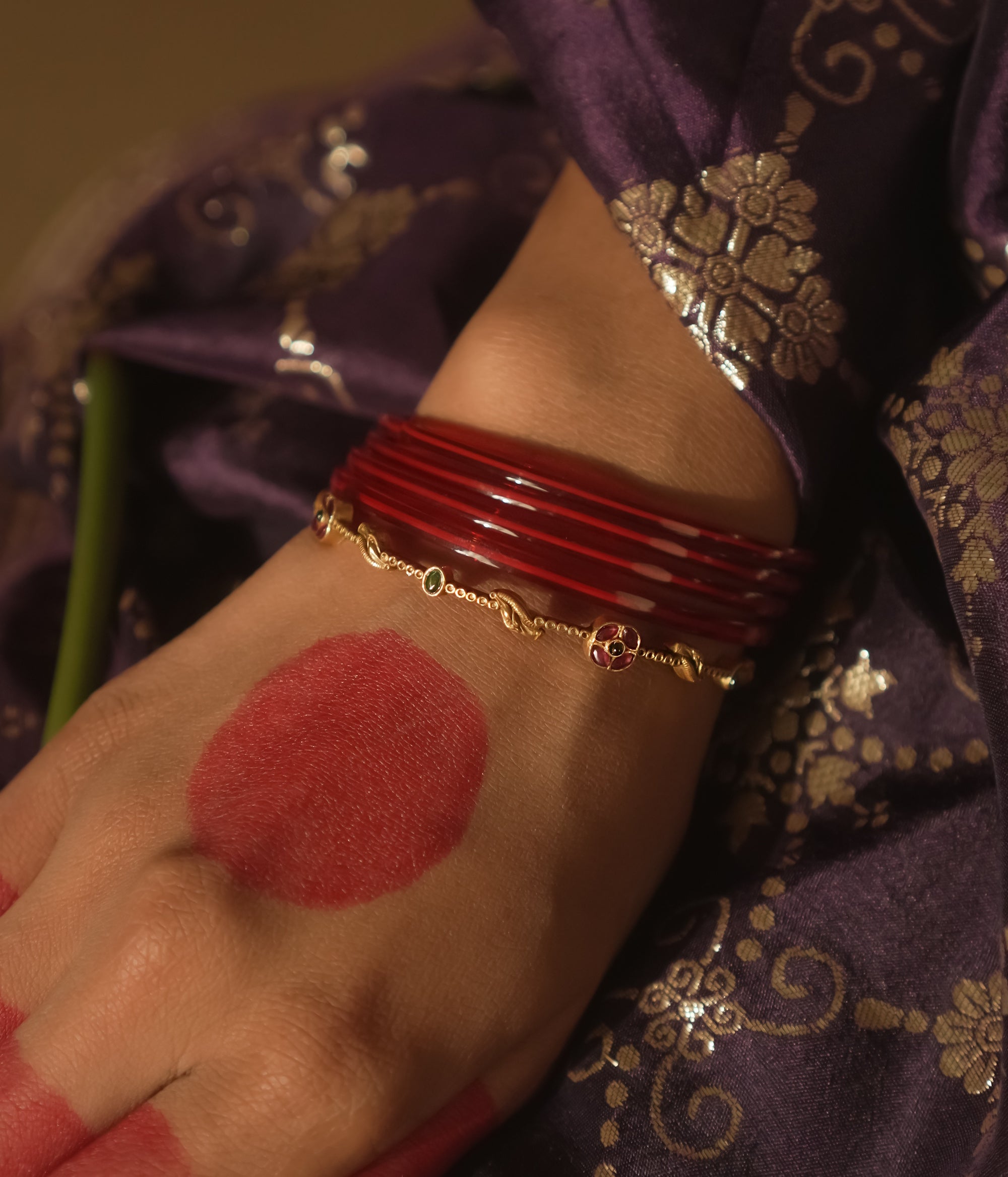 Hand with red bangles and a red dot, wearing a purple saree with gold patterns.