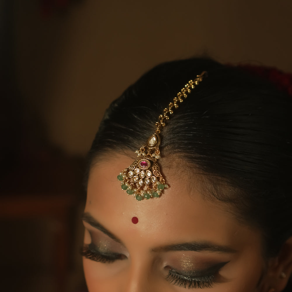 Gold maangtika with a central red gem, emerald beads, and a delicate chain.