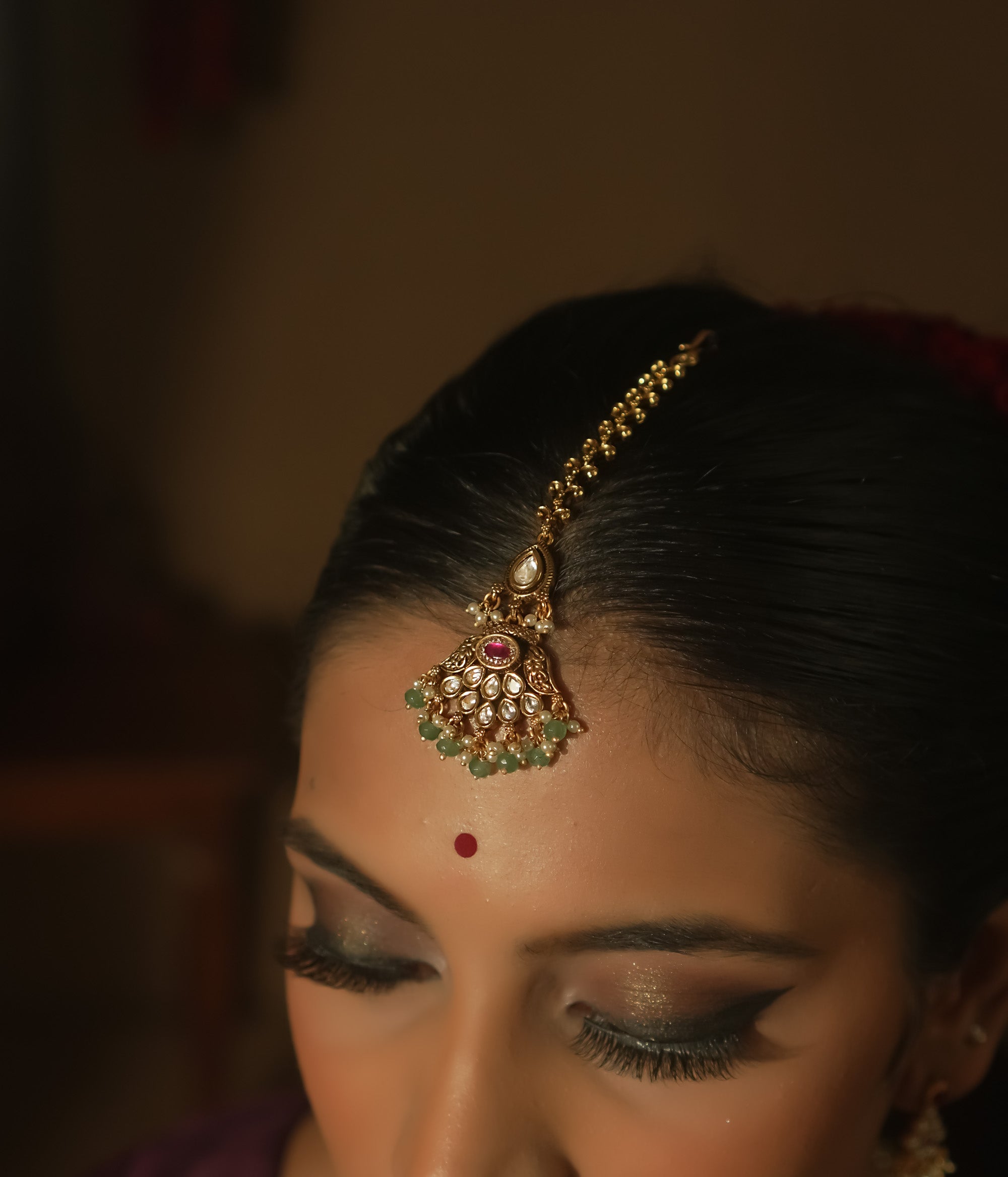 Gold maangtika with a central red gem, emerald beads, and a delicate chain.