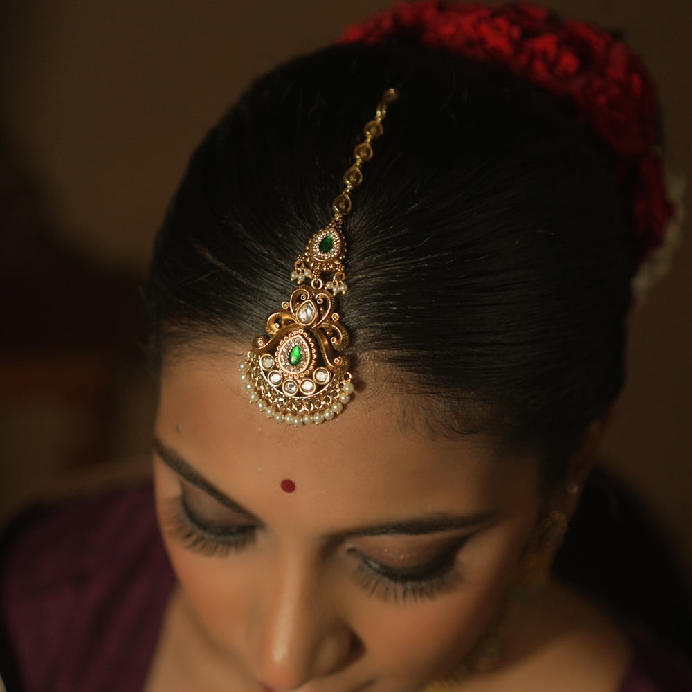 Gold maangtika with a central green teardrop gemstone, pearls, and red floral motifs.