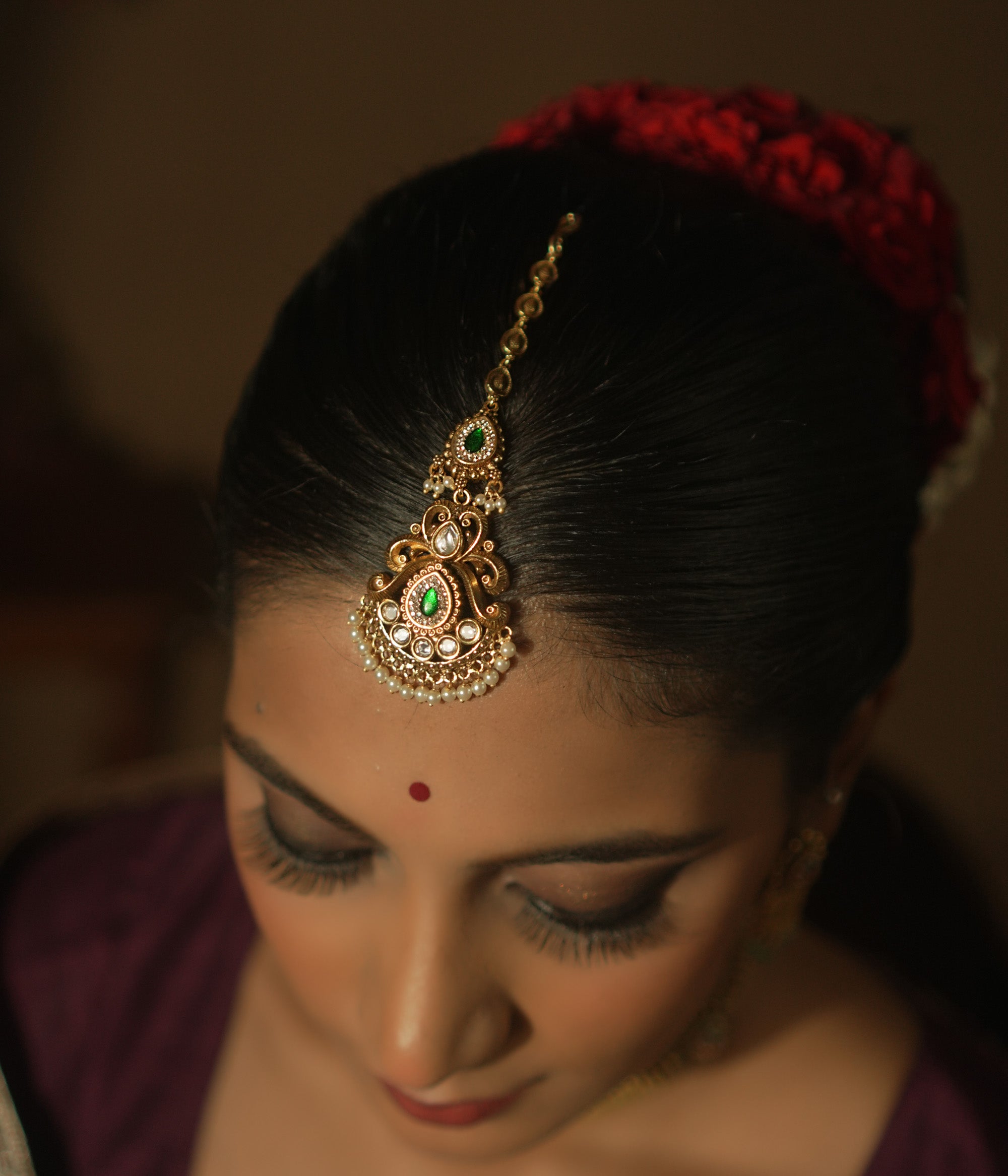 Gold maangtika with a central green teardrop gemstone, pearls, and red floral motifs.
