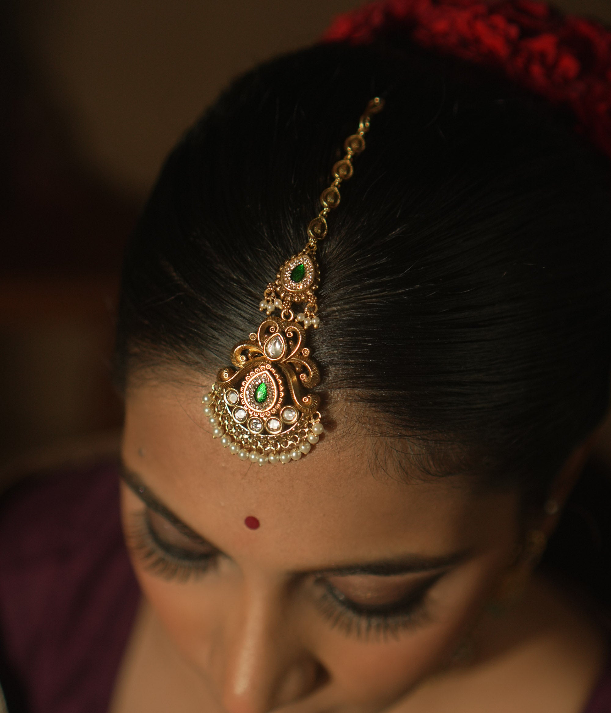 Gold maangtika with a central green teardrop gemstone, pearls, and red floral motifs.