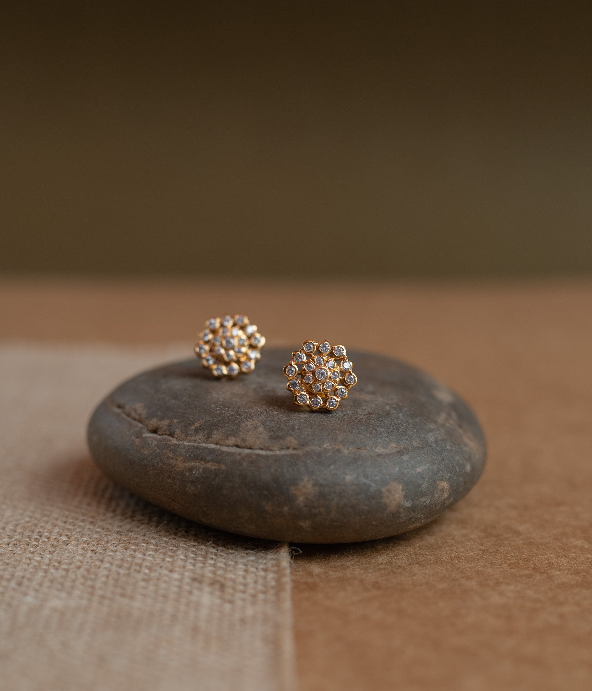 Gold floral stud earrings covered in clear Swarovski stones, displayed on a textured fabric.
