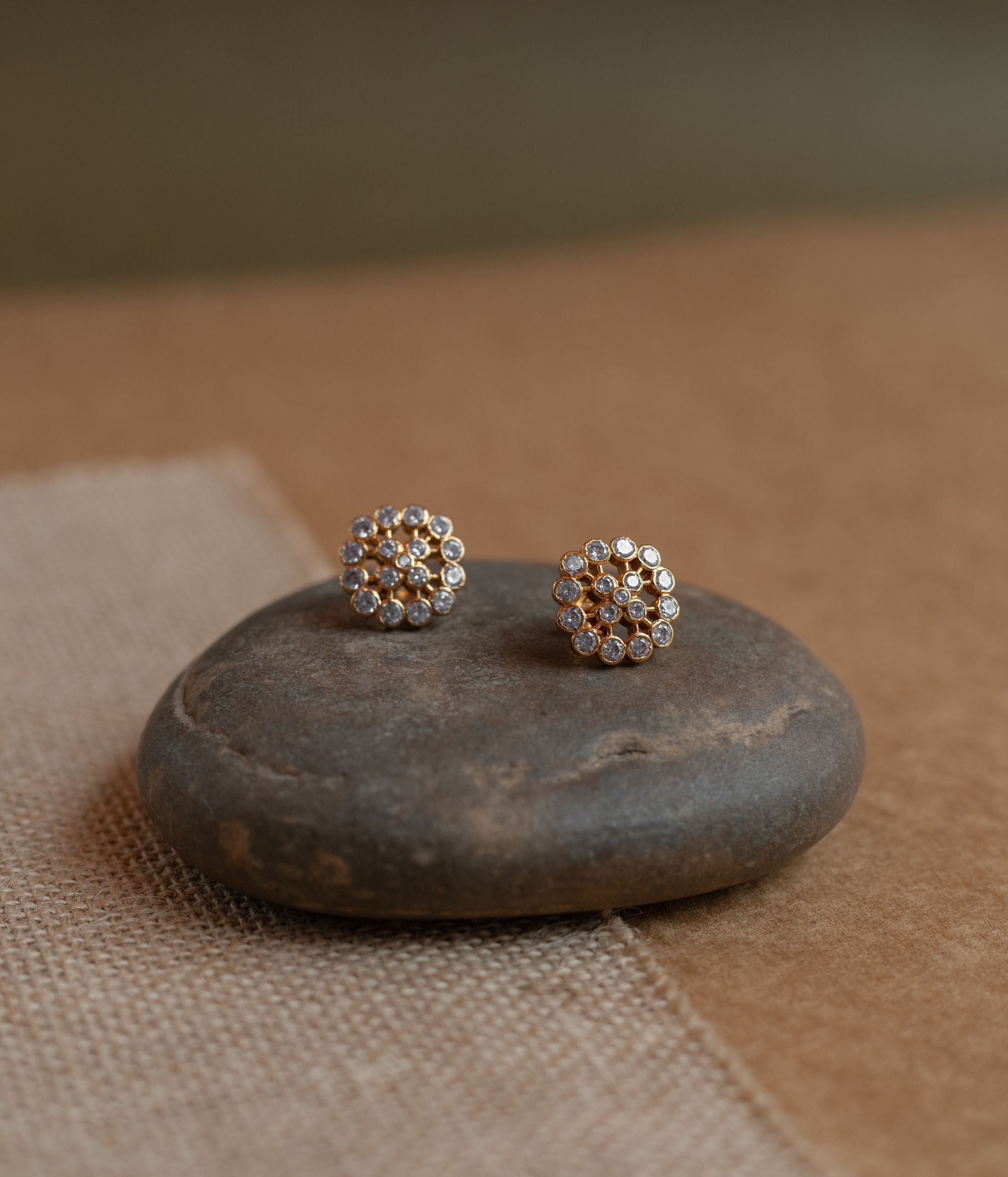 Gold floral stud earrings covered in clear Swarovski stones, displayed on a textured fabric.