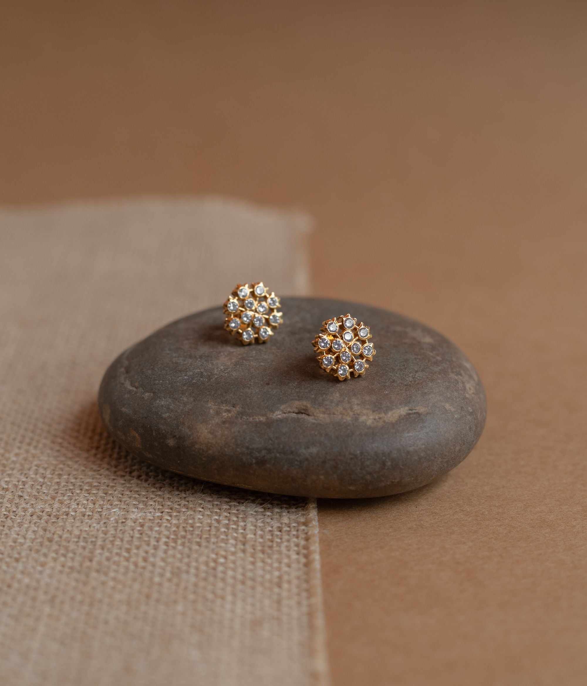 Gold floral stud earrings covered in clear Swarovski stones, displayed on a textured fabric.