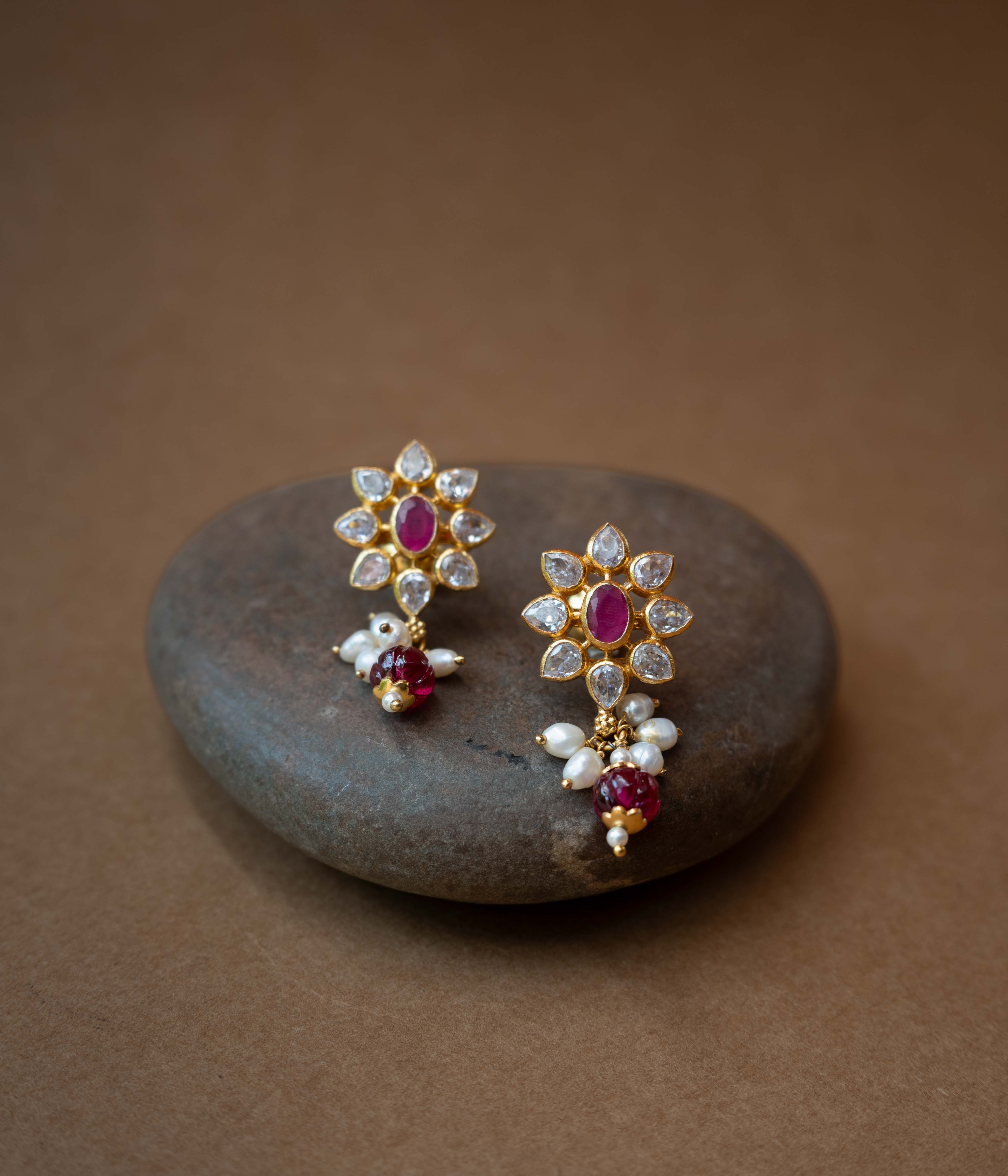 A pair of intricate floral earrings with a central red stone, surrounded by clear stones, and dangling pearls.