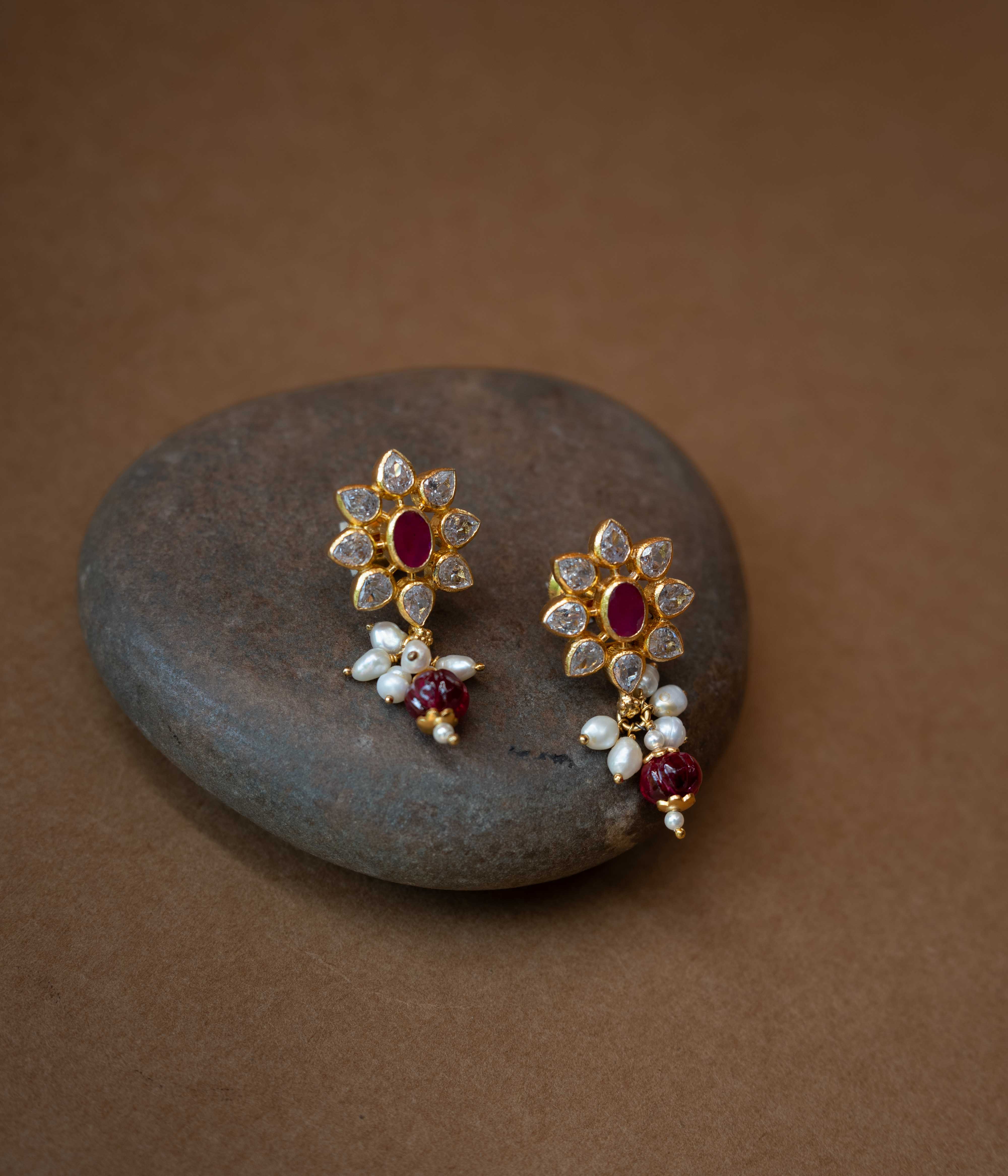 A pair of intricate floral earrings with a central red stone, surrounded by clear stones, and dangling pearls.