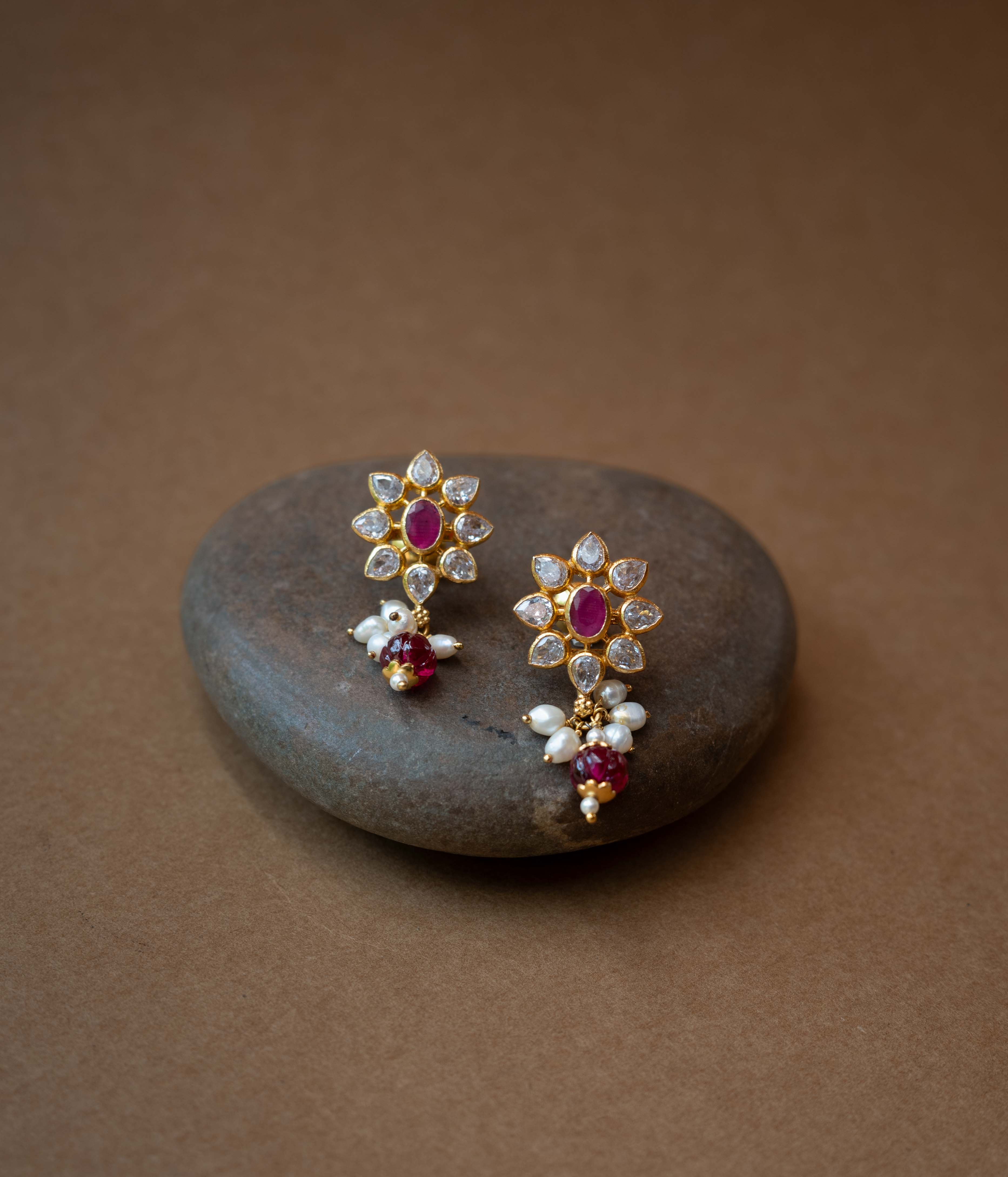 A pair of intricate floral earrings with a central red stone, surrounded by clear stones, and dangling pearls.